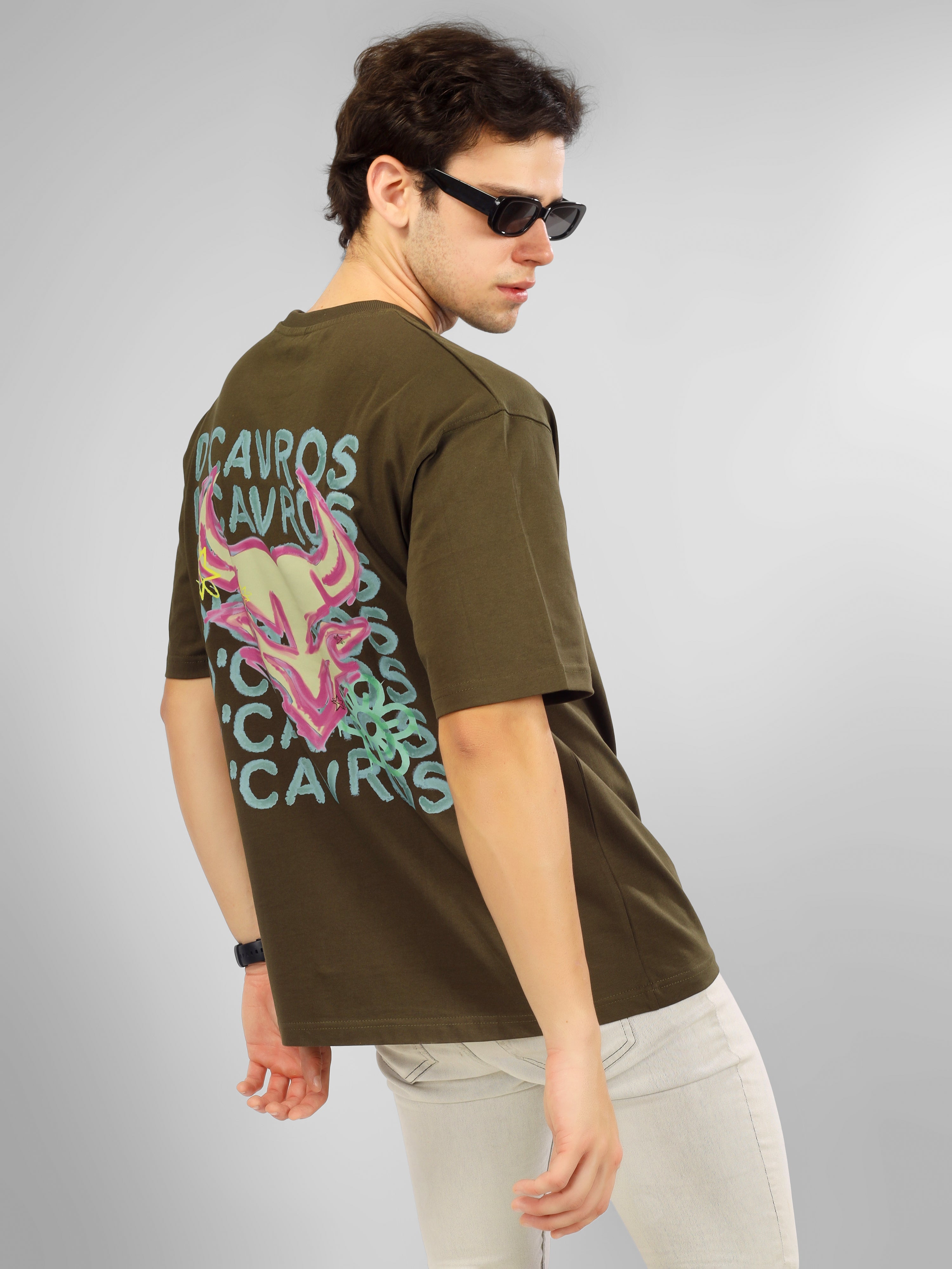 Man wearing olive green oversized D'Cavros T-shirt featuring a vibrant bull graphic and repeated D'Cavros logo text on the back, styled with beige jeans and sneakers.