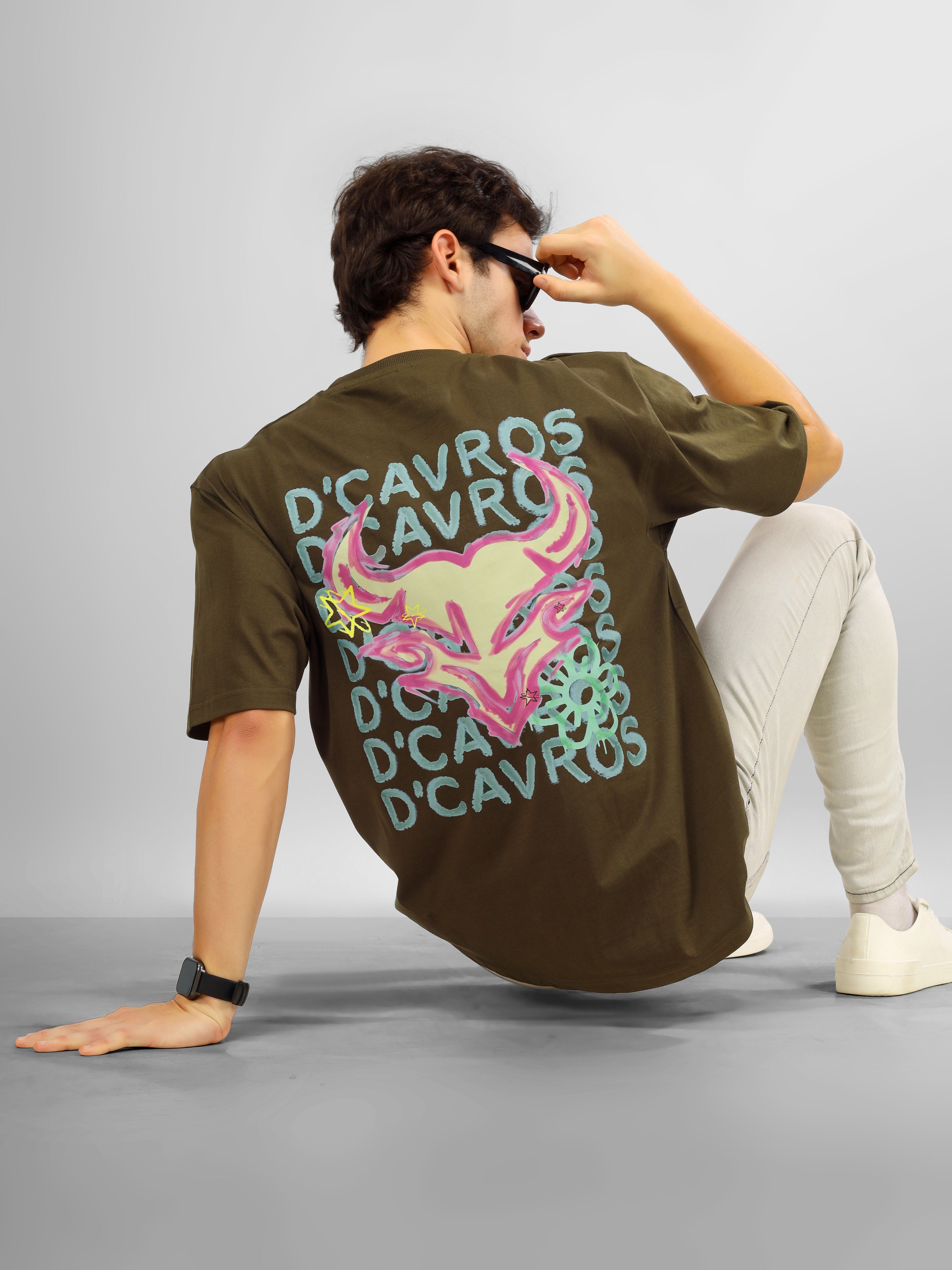 Man wearing olive green oversized D'Cavros T-shirt featuring a vibrant bull graphic and repeated D'Cavros logo text on the back, styled with beige jeans and sneakers.