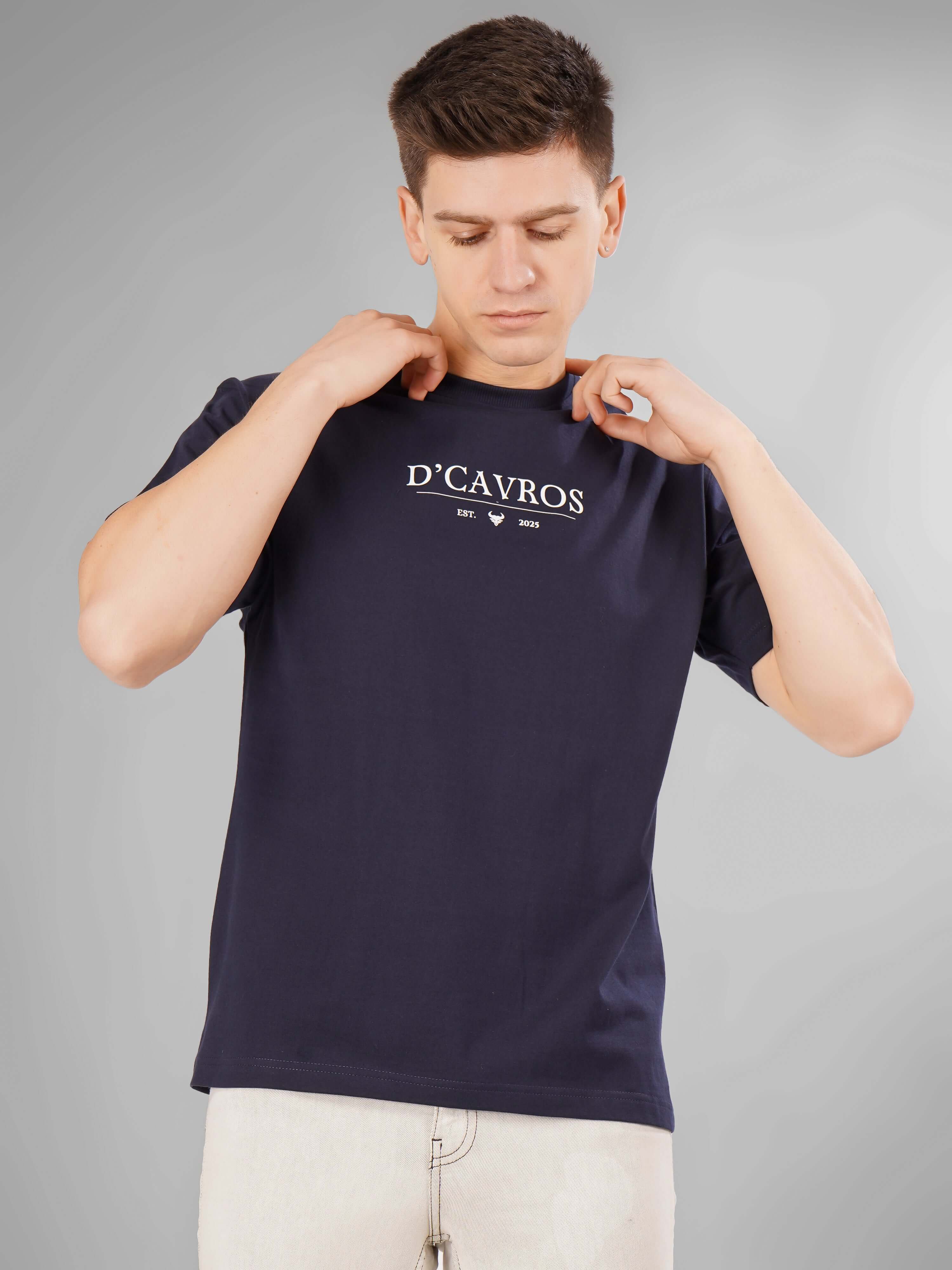 Man wearing Solid Logo for Men a navy blue D'Cavros T-shirt with a bold white printed logo on the chest, paired with light gray jeans, posing confidently against a plain grey background.