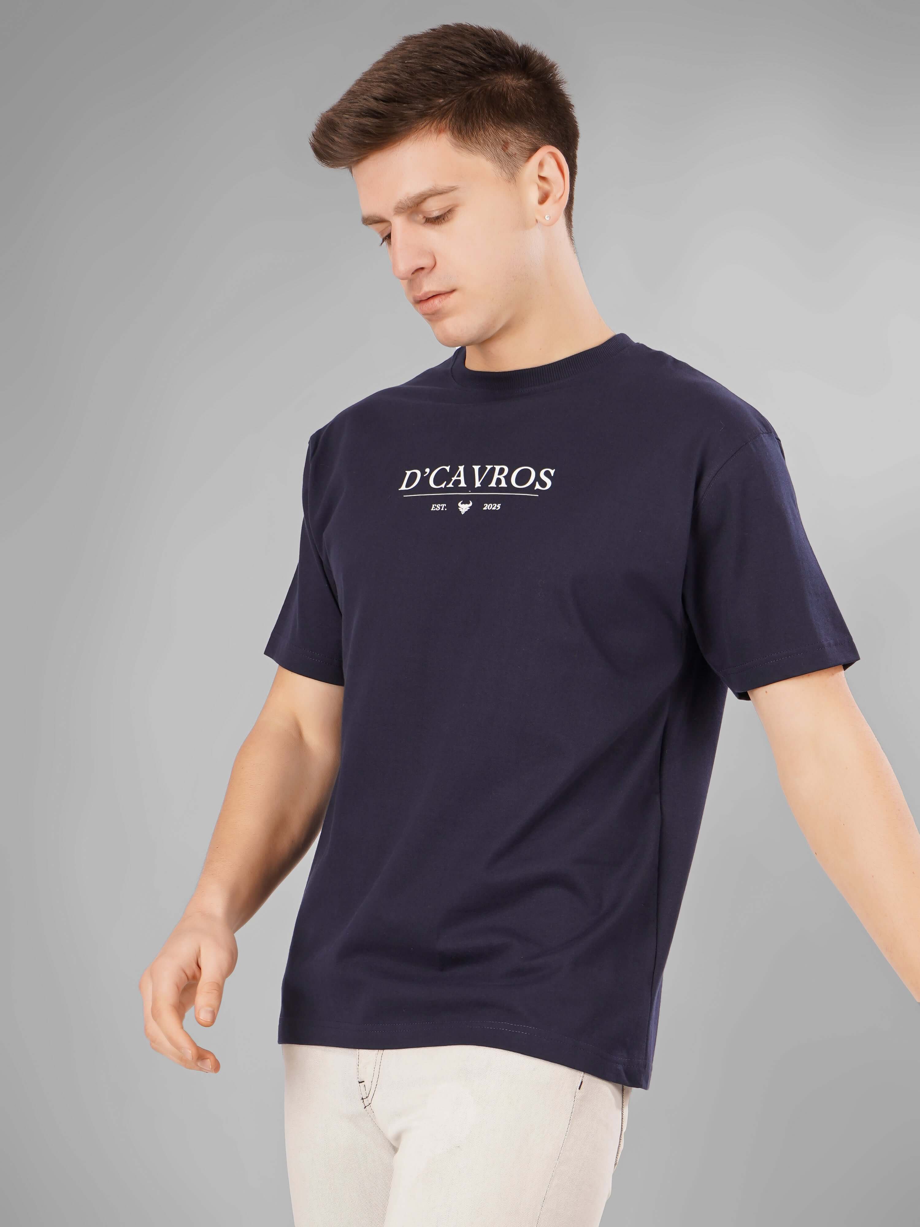 Man wearing Solid Logo for Men a navy blue D'Cavros T-shirt with a bold white printed logo on the chest, paired with light gray jeans, posing confidently against a plain grey background.