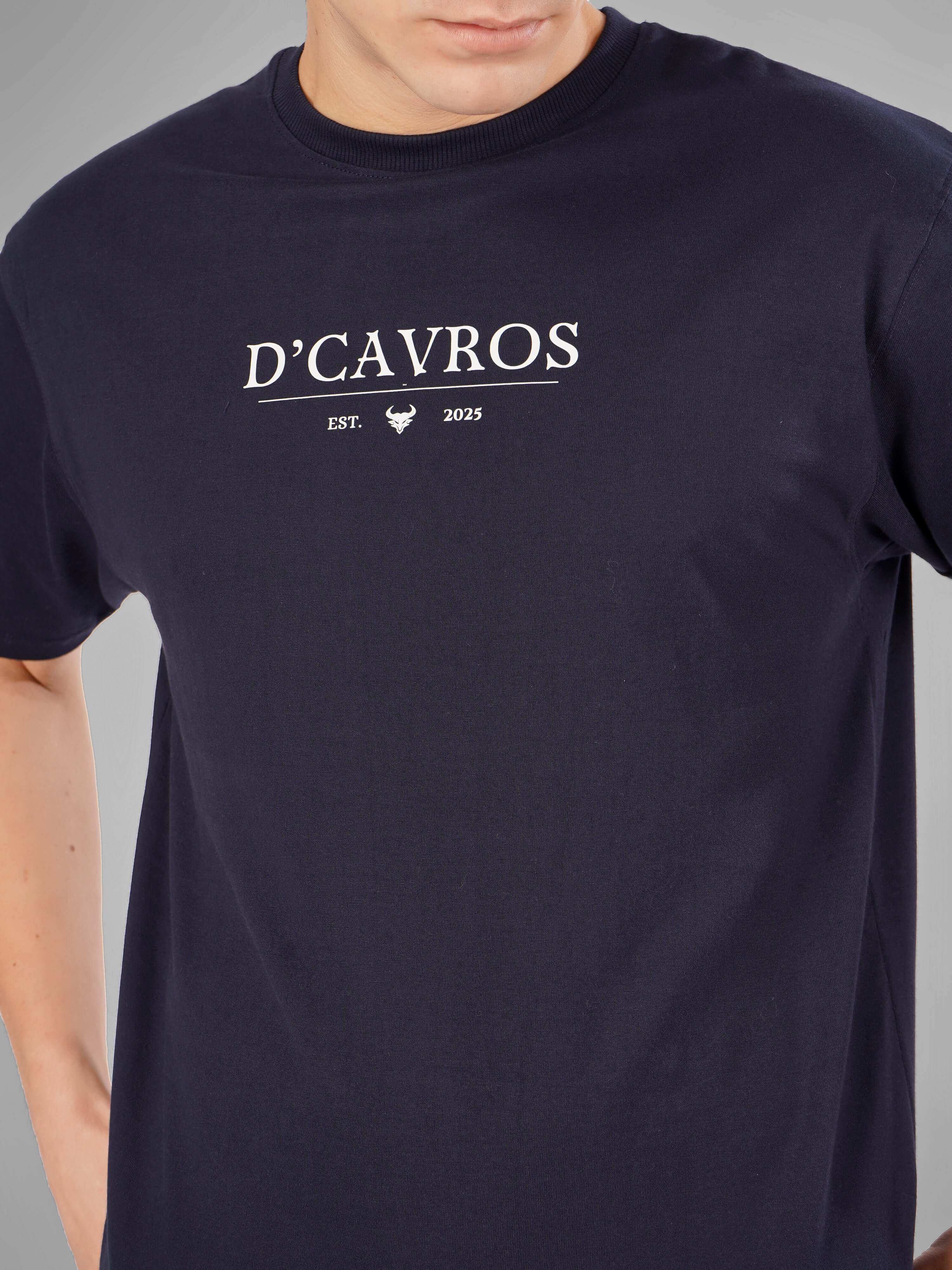 Man wearing Solid Logo for Men a navy blue D'Cavros T-shirt with a bold white printed logo on the chest, paired with light gray jeans, posing confidently against a plain grey background.