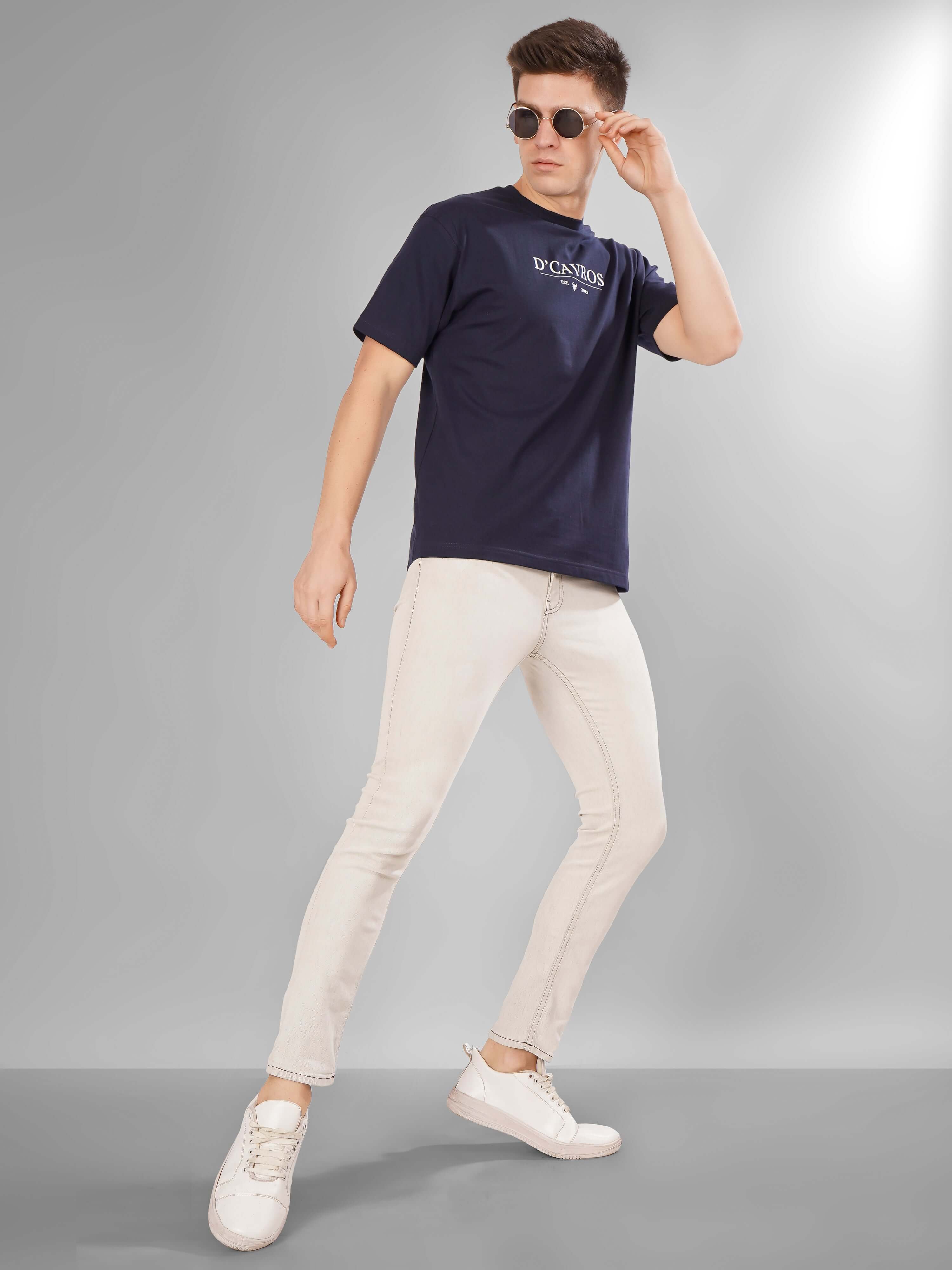 Man wearing Solid Logo for Men a navy blue D'Cavros T-shirt with a bold white printed logo on the chest, paired with light gray jeans, posing confidently against a plain grey background.