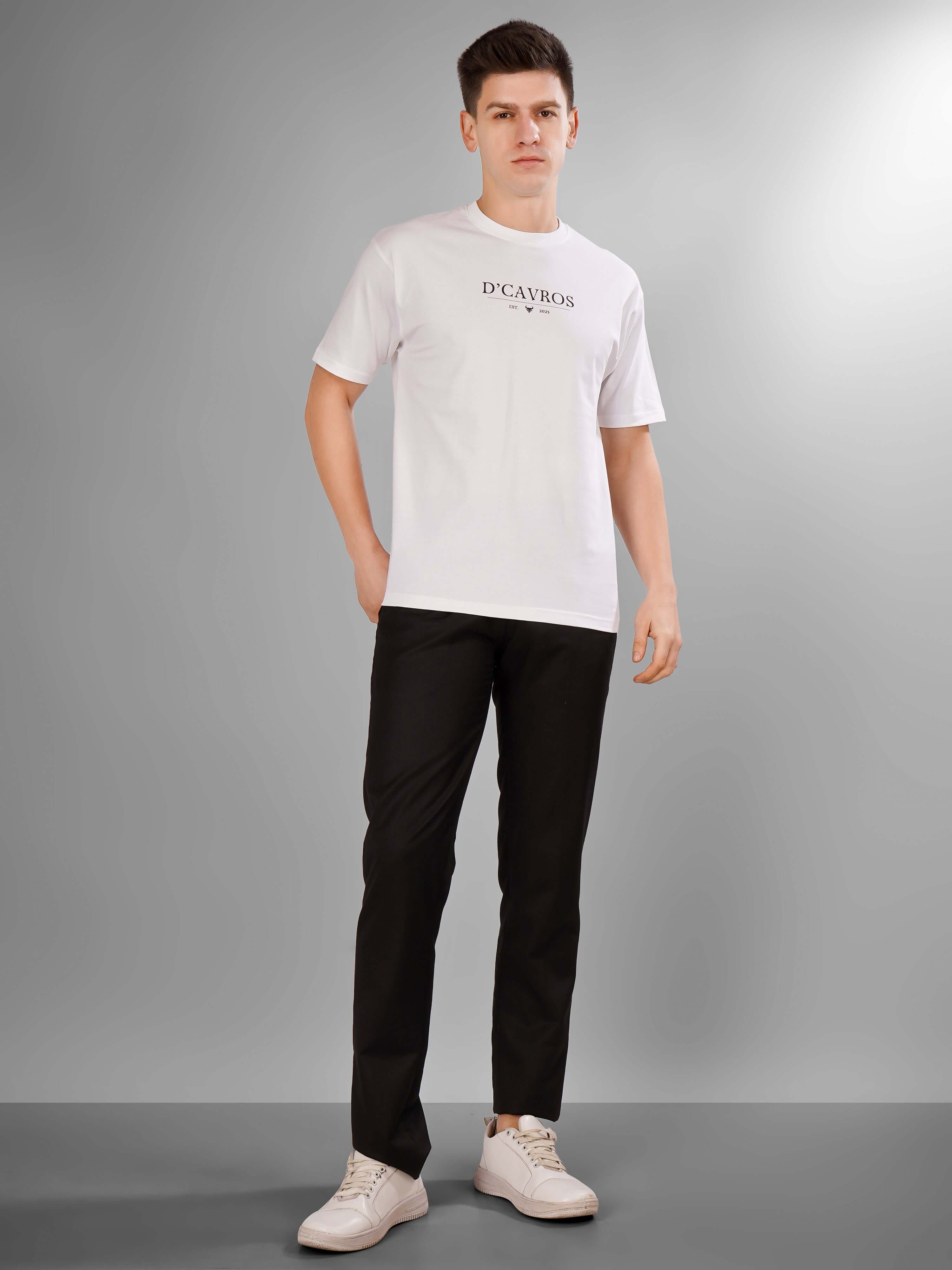 Man wearing Solid Logo for Men a white D'Cavros T-shirt with a bold black printed logo on the chest, paired with black pants and white sneakers, standing against a plain grey background.