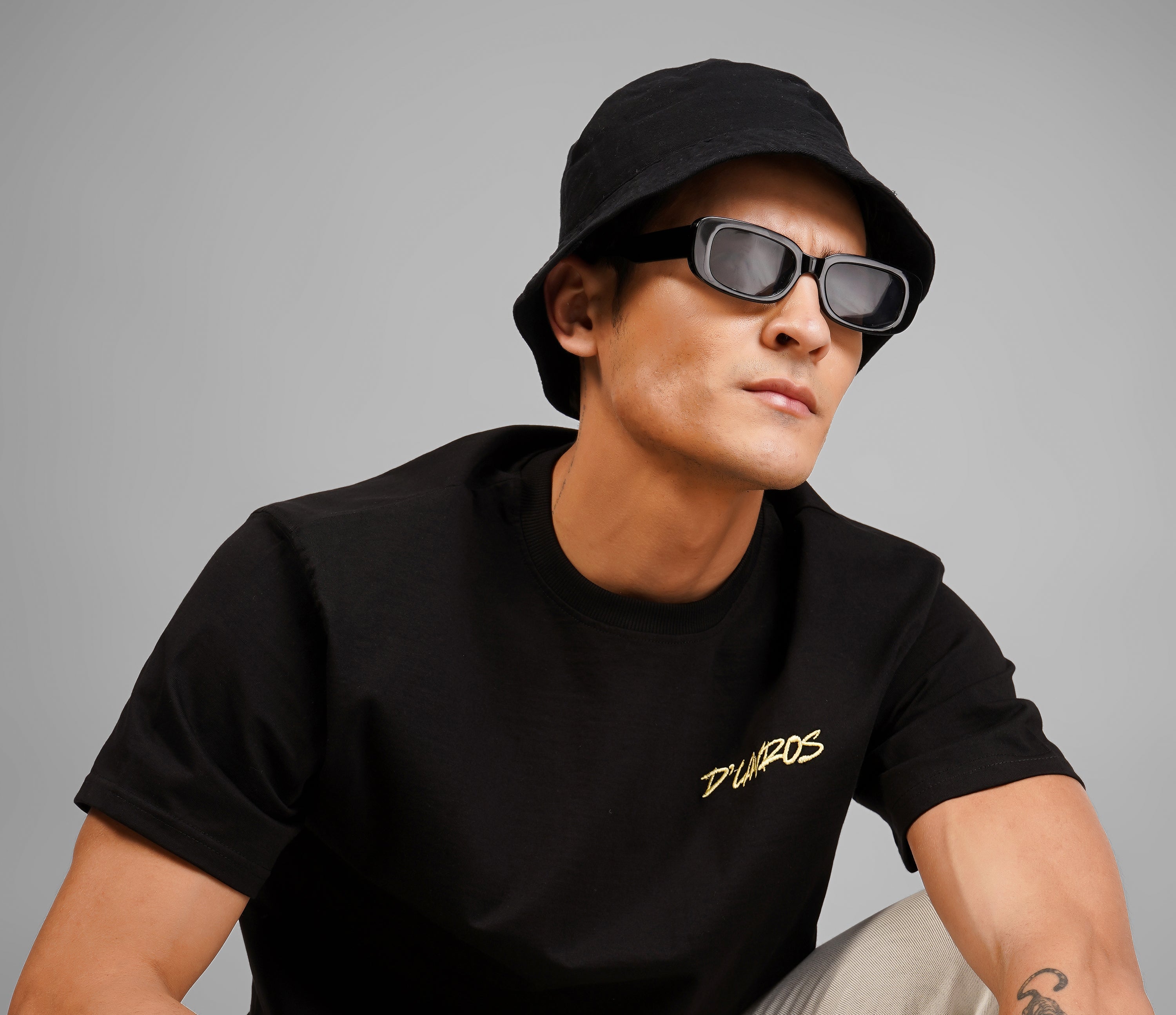 Man wearing a black t-shirt with a logo, beige pants, and a black cap on a gray background