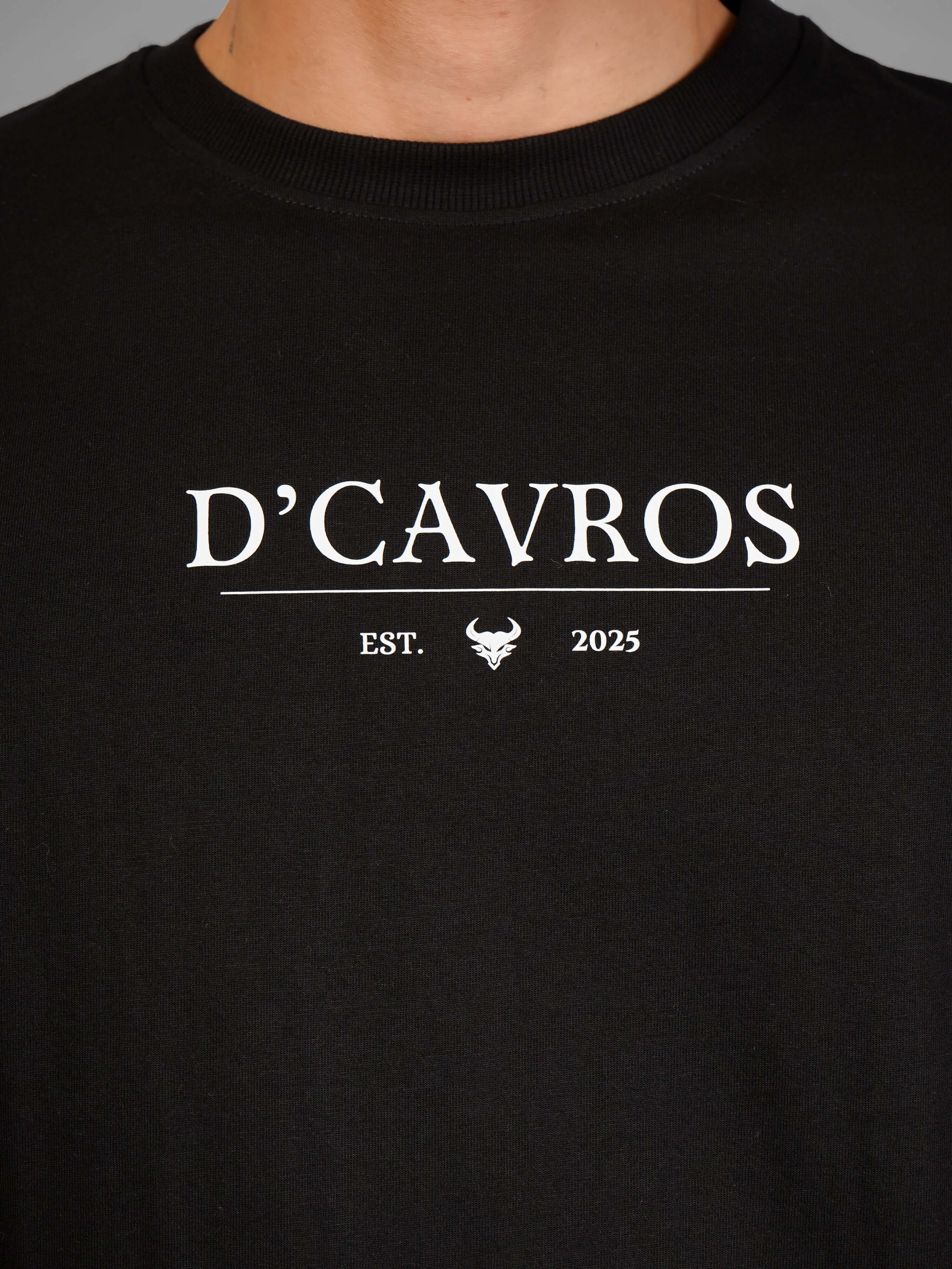 Man wearing Solid Logo for Men a black D'Cavros T-shirt with a printed dcavros logo on the chest, paired with light gray jeans and black sunglasses, posing against a plain grey background.