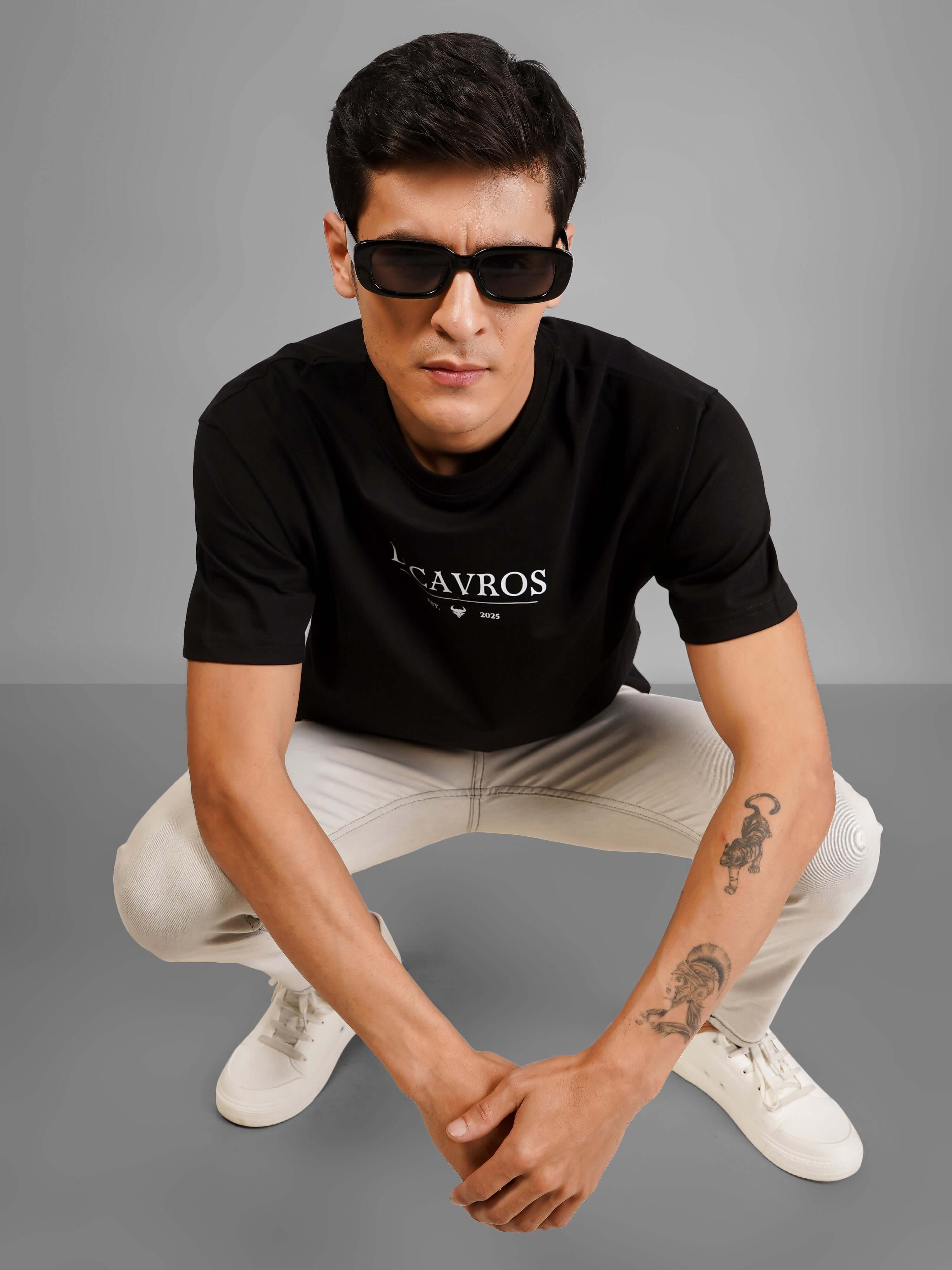 Man wearing Solid Logo for Men a black D'Cavros T-shirt with a printed dcavros logo on the chest, paired with light gray jeans and black sunglasses, posing against a plain grey background.