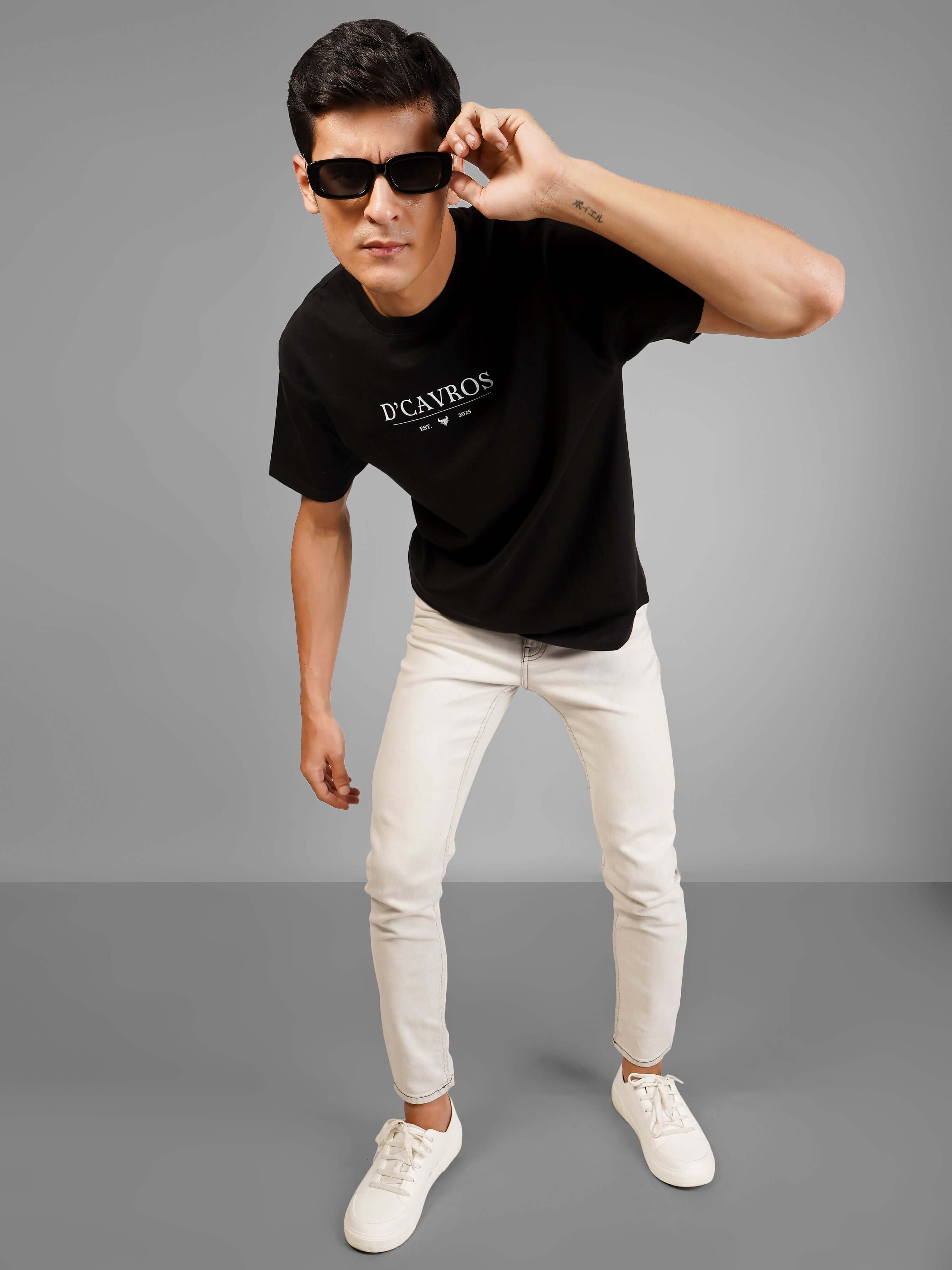 Man wearing Solid Logo for Men a black D'Cavros T-shirt with a printed dcavros logo on the chest, paired with light gray jeans and black sunglasses, posing against a plain grey background.
