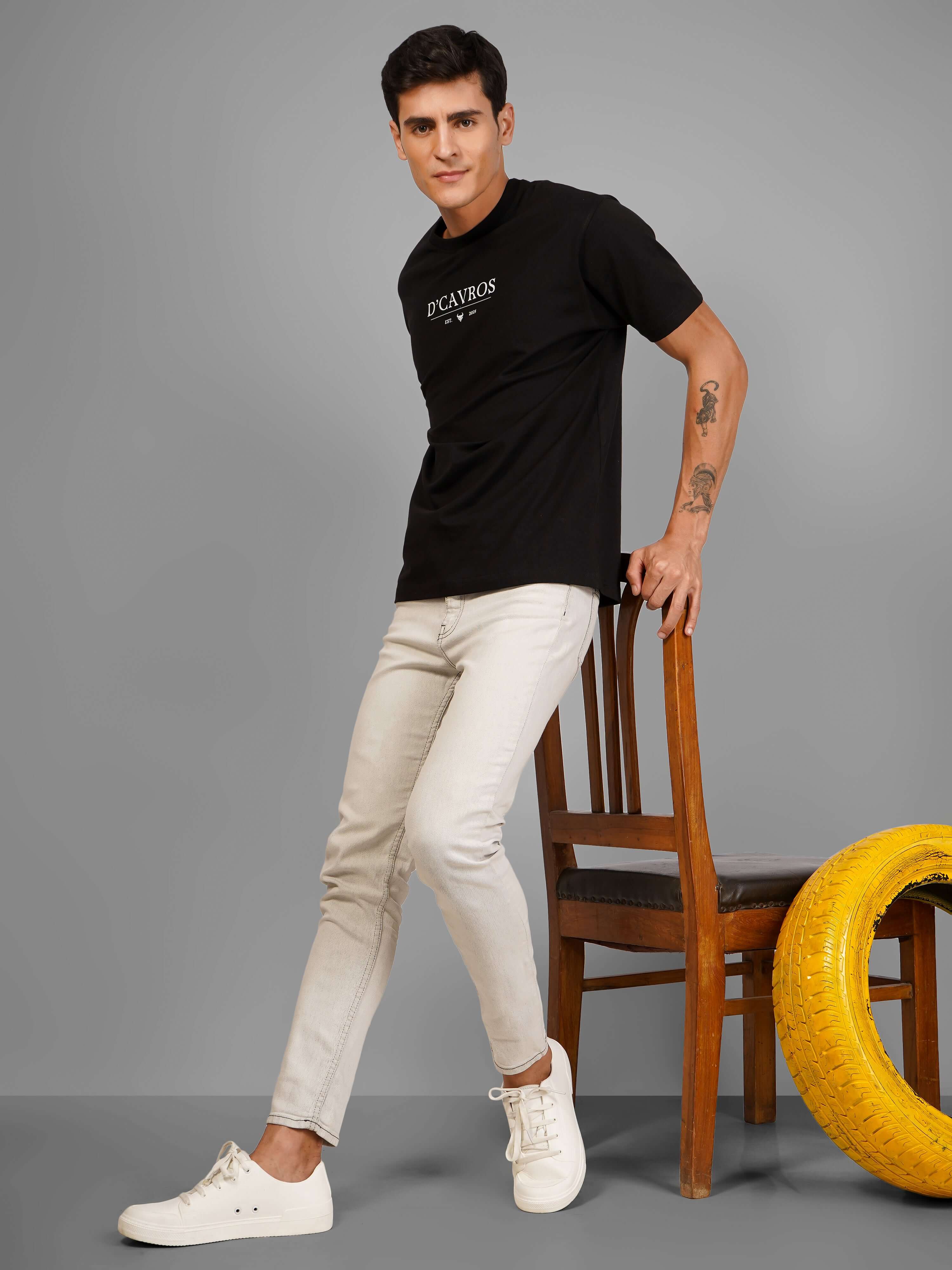 Man wearing Solid Logo for Men a black D'Cavros T-shirt with a printed dcavros logo on the chest, paired with light gray jeans and black sunglasses, posing against a plain grey background.