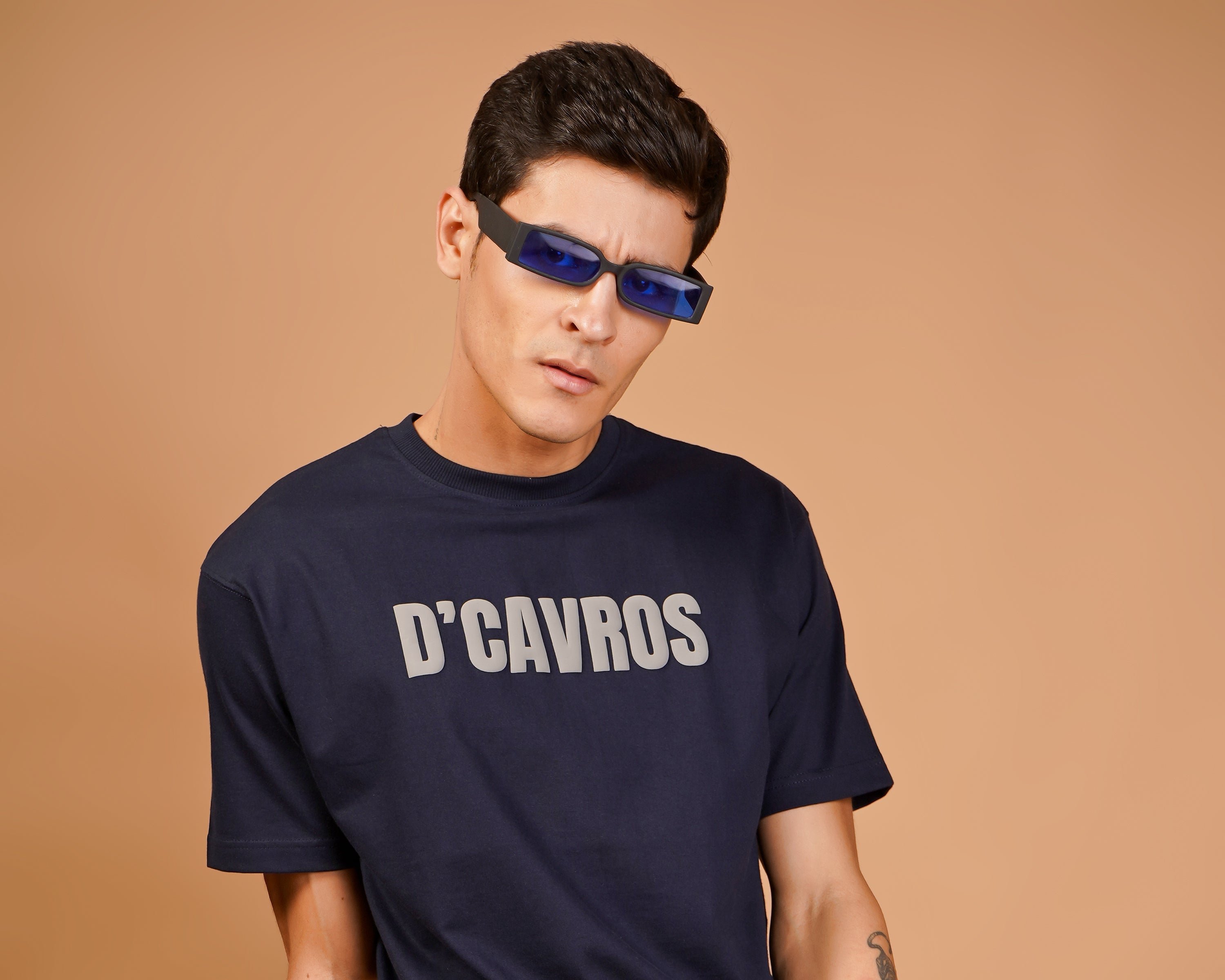 Man wearing a navy blue t-shirt with 'D'CAVROS' text, white pants, and sunglasses on a brown background.