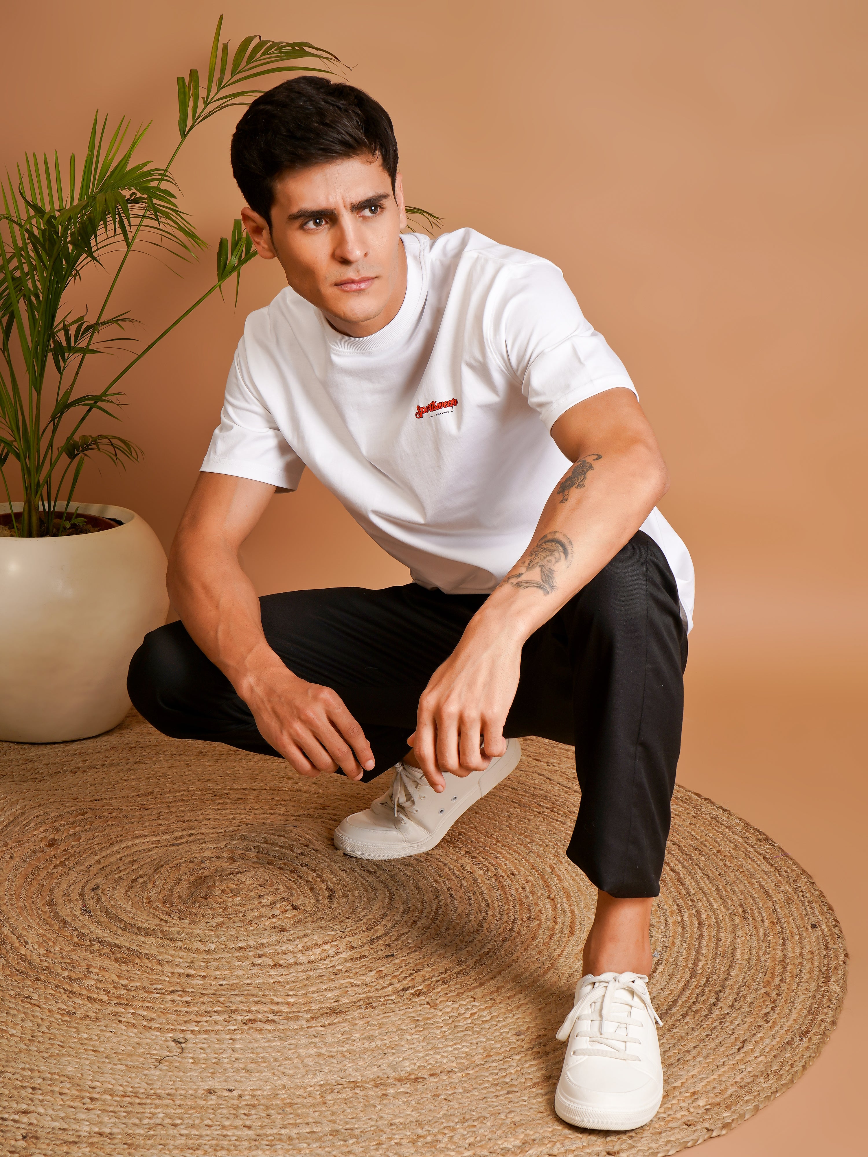 Man squatting in a Sports Car Oversized T-Shirt against a neutral background with indoor plants.