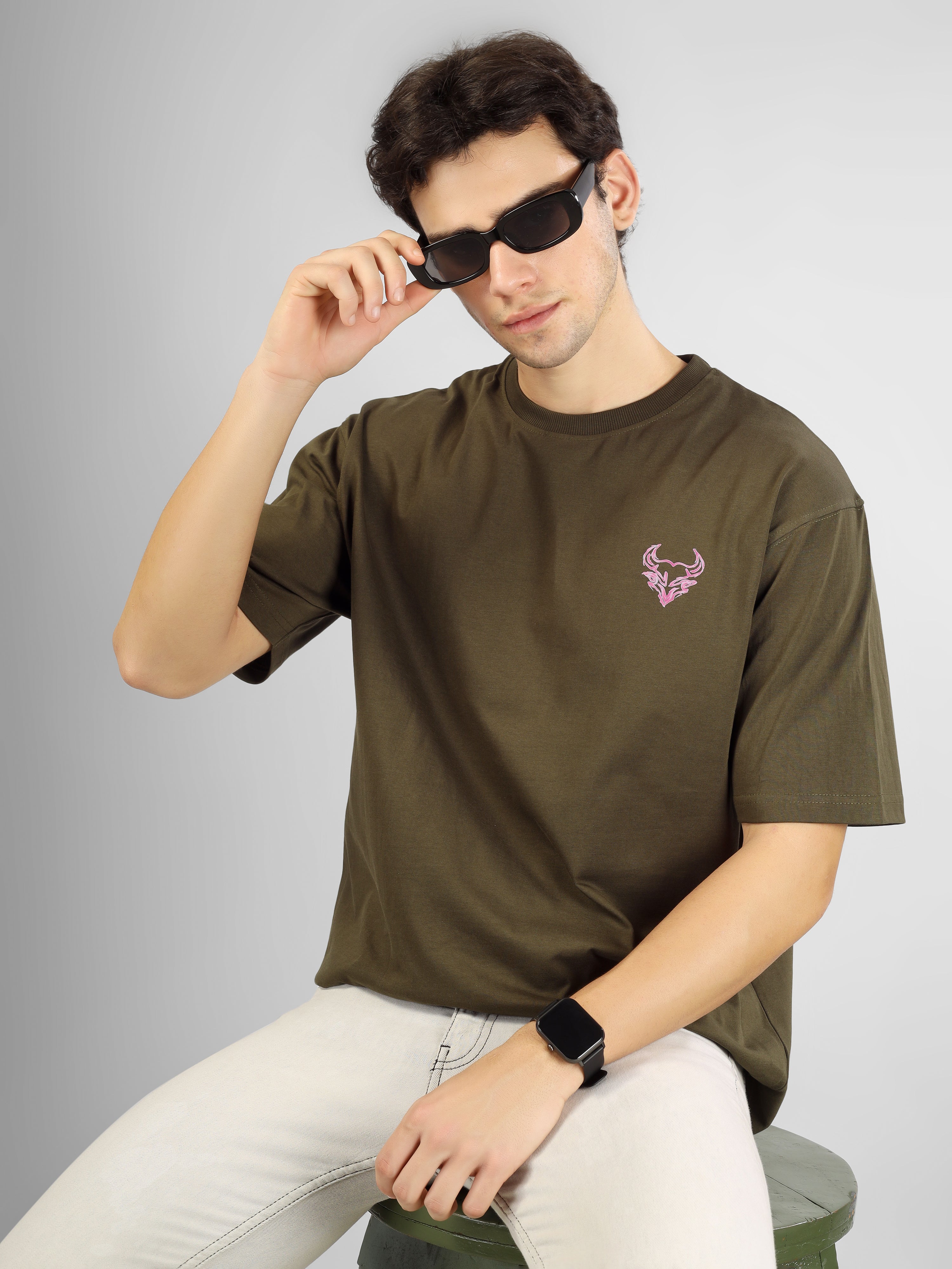 Man wearing olive green oversized D'Cavros T-shirt featuring a vibrant bull graphic and repeated D'Cavros logo text on the back, styled with beige jeans and sneakers.