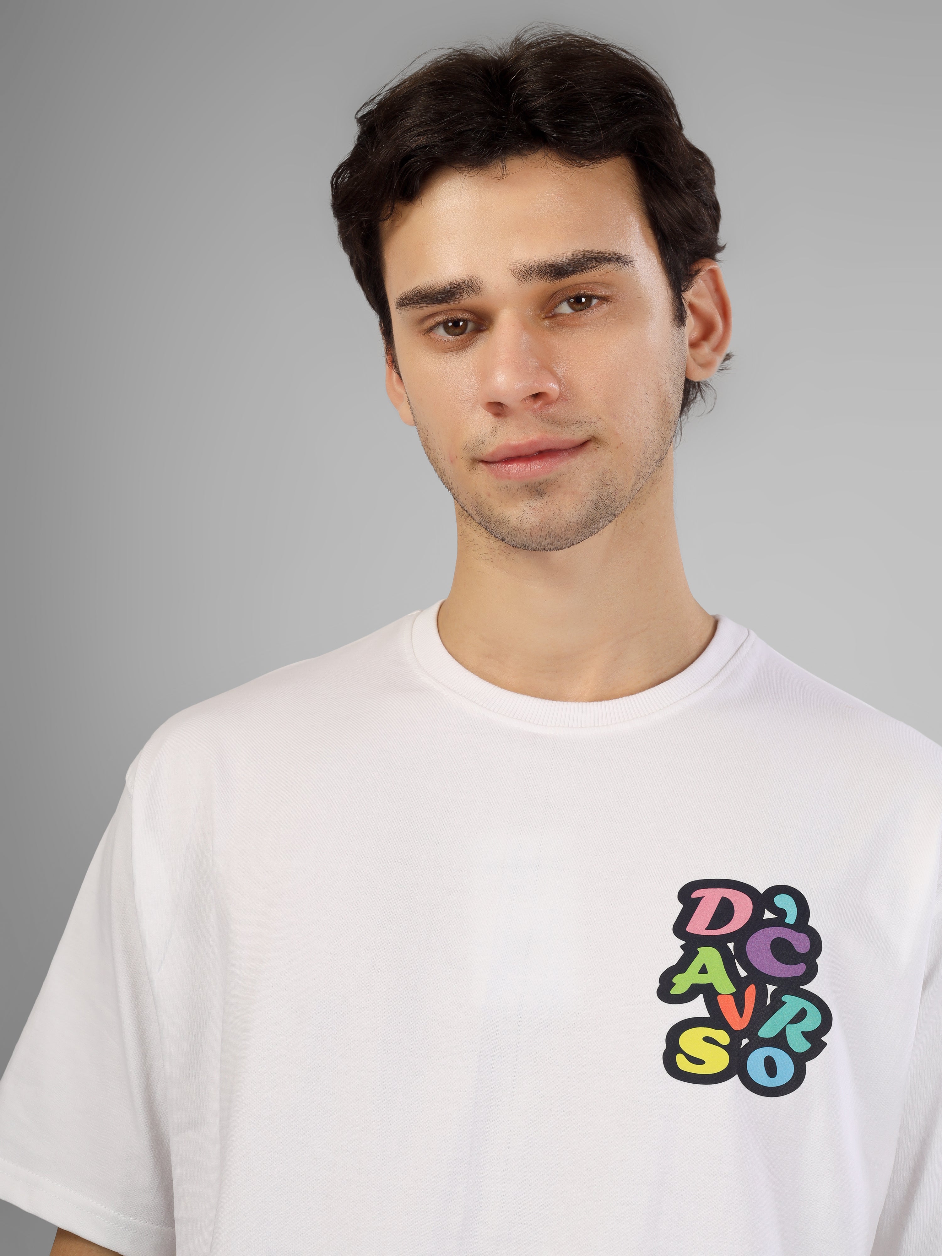 Male model wearing a white oversized dcavros T-shirt with a multicolor graphic logo on the left chest, styled with black pants and sunglasses.