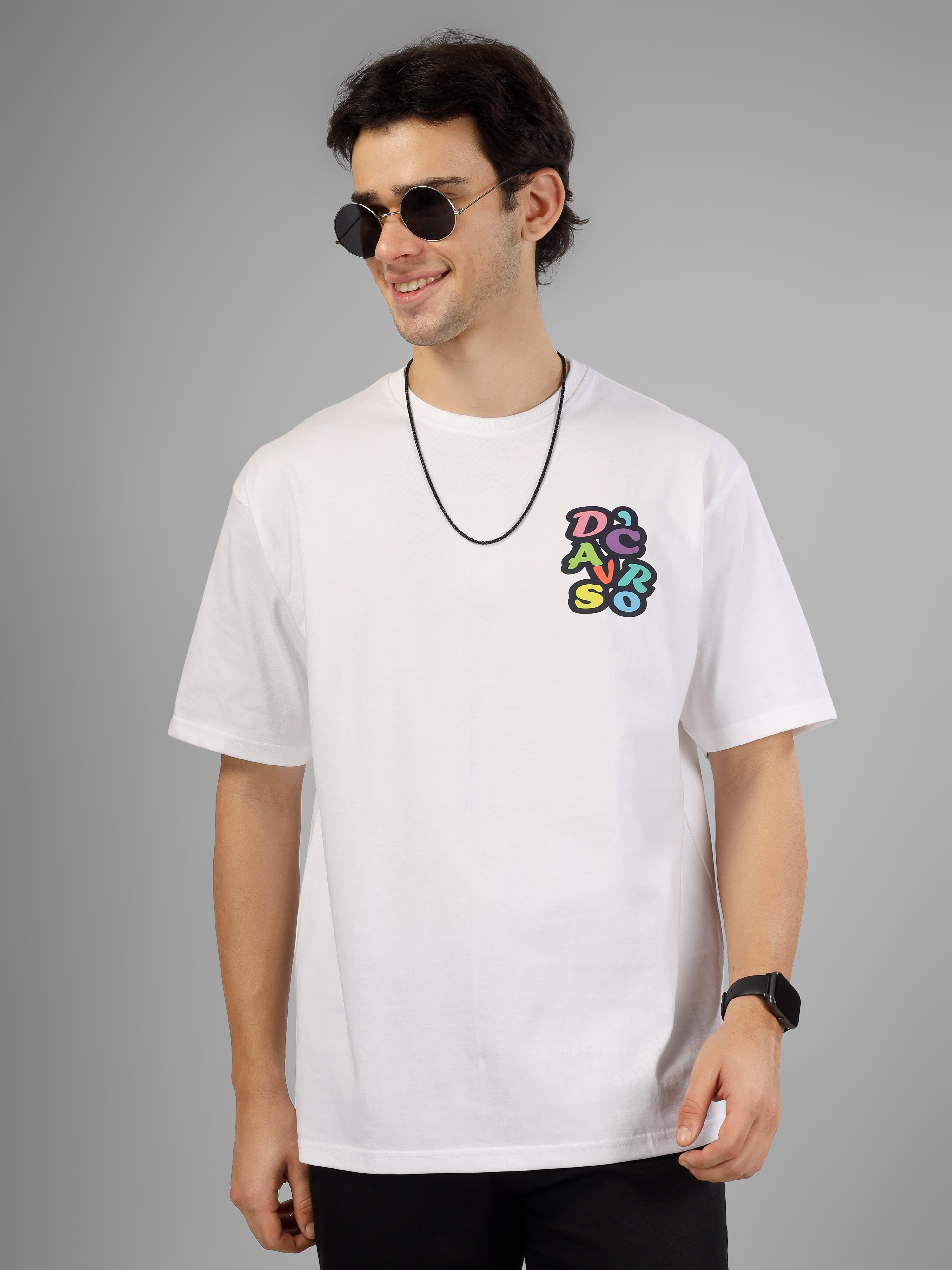 Male model wearing a white oversized dcavros T-shirt with a multicolor graphic logo on the left chest, styled with black pants and sunglasses.