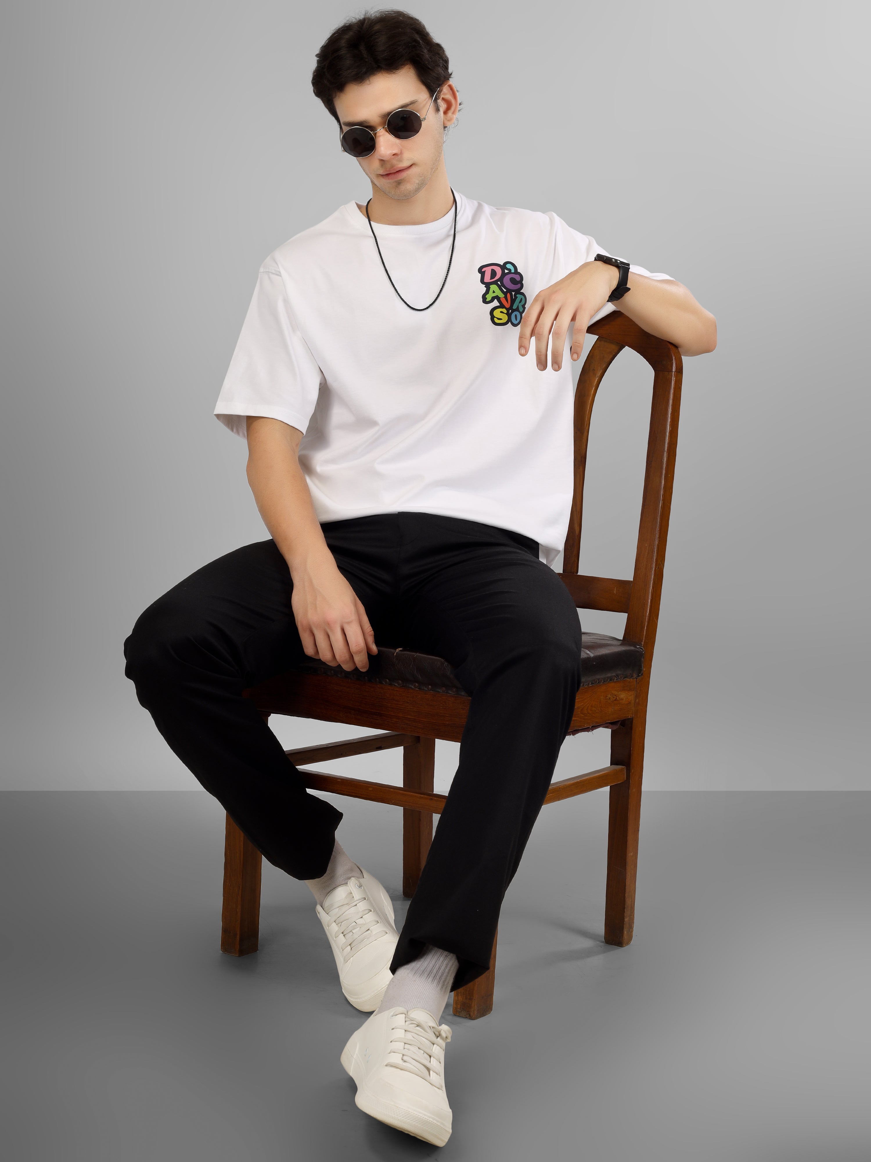 Male model wearing a white oversized dcavros T-shirt with a multicolor graphic logo on the left chest, styled with black pants and sunglasses.