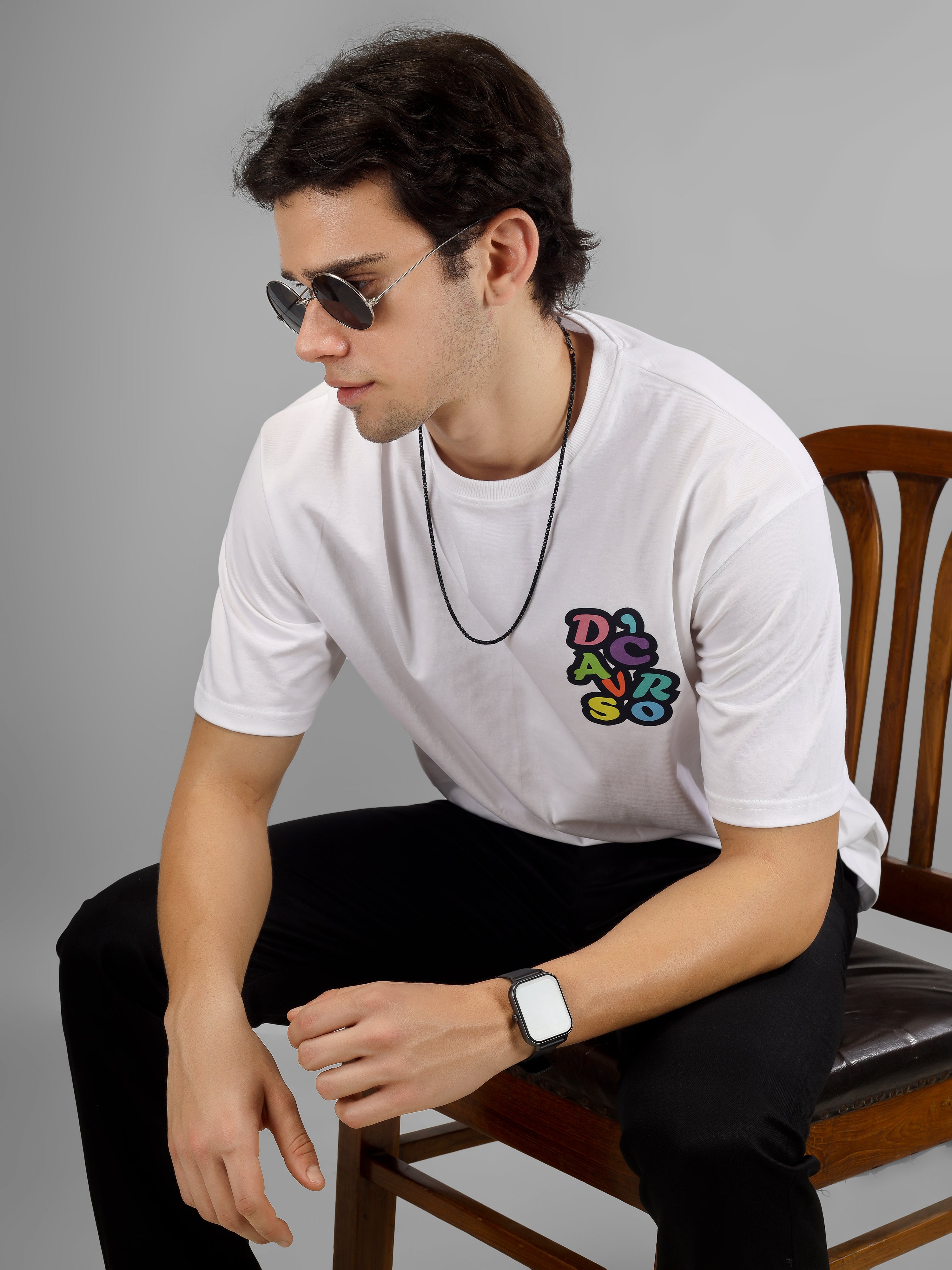 Male model wearing a white oversized dcavros T-shirt with a multicolor graphic logo on the left chest, styled with black pants and sunglasses.