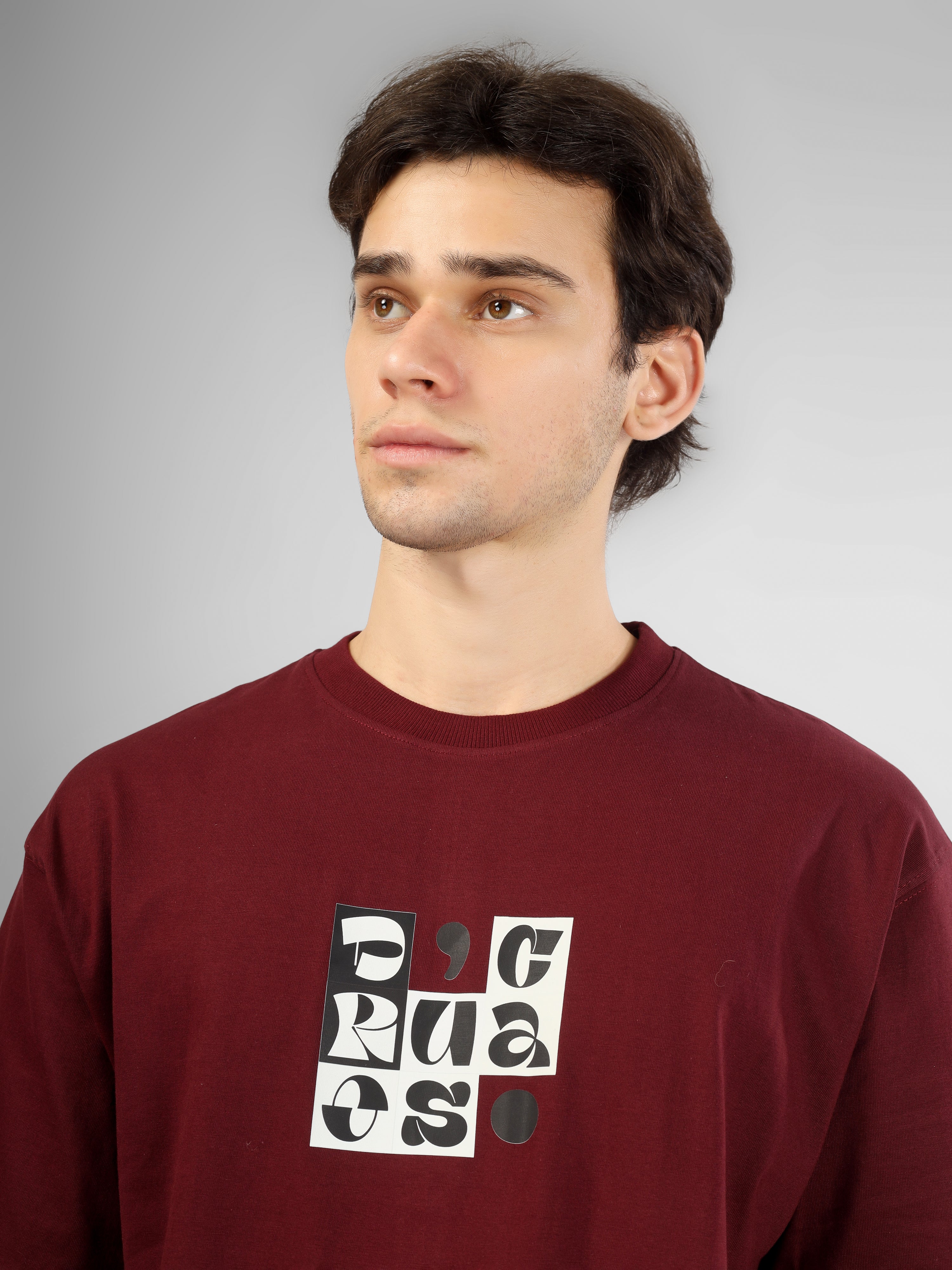 Male model wearing a maroon dcavros T-shirt featuring a black-and-white abstract center logo print, paired with beige pants.