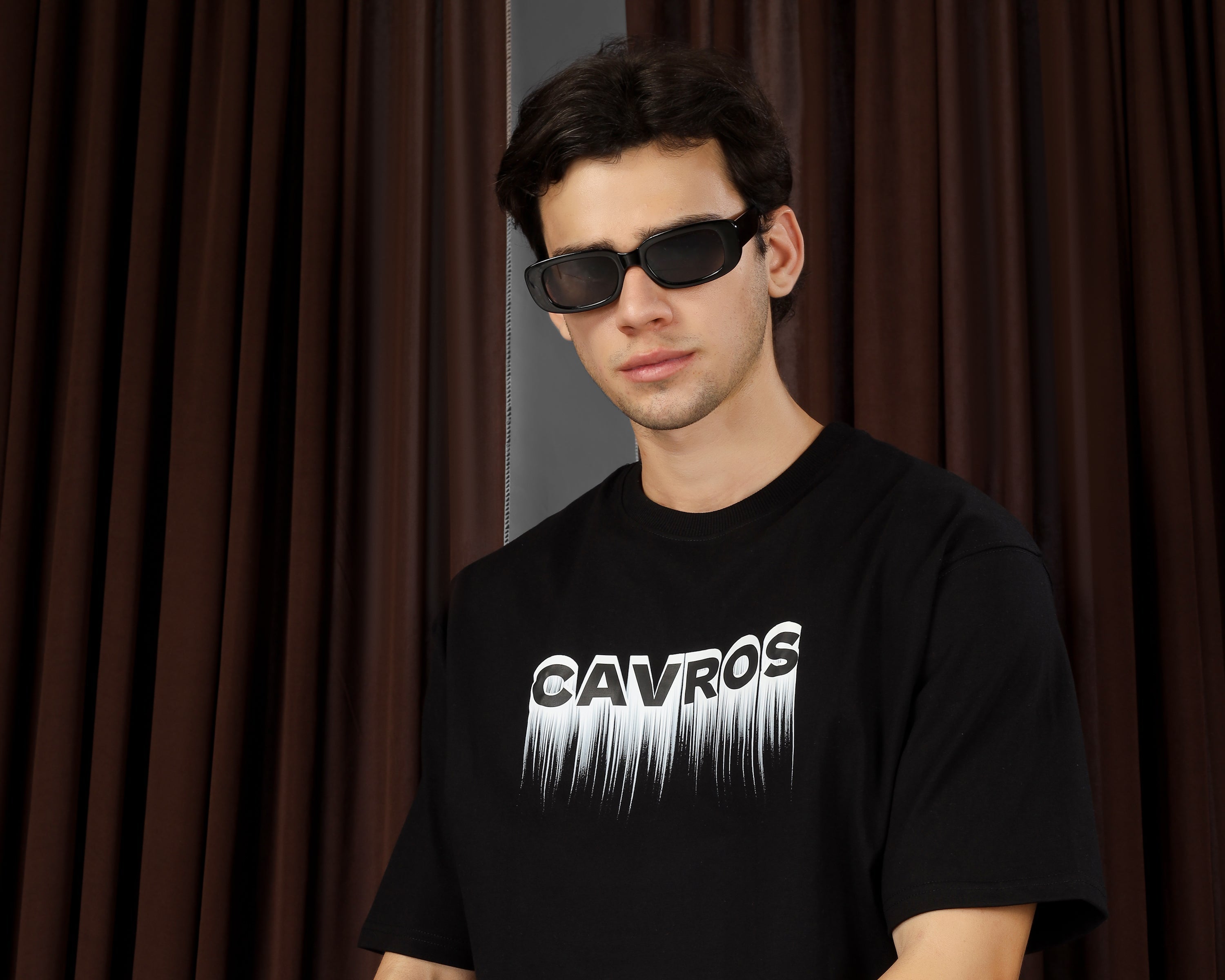 Person wearing a black t-shirt with 'CAVROS' logo sitting against a brown curtain.