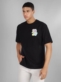 Male model wearing a black oversized dcavros T-shirt with a vibrant multicolor logo on the left chest, paired with white jeans.