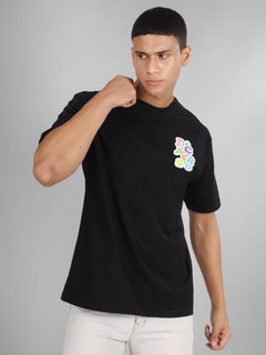 Male model wearing a black oversized dcavros T-shirt with a vibrant multicolor logo on the left chest, paired with white jeans.