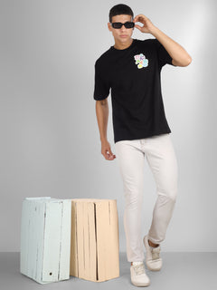 Male model wearing a black oversized dcavros T-shirt with a vibrant multicolor logo on the left chest, paired with white jeans.