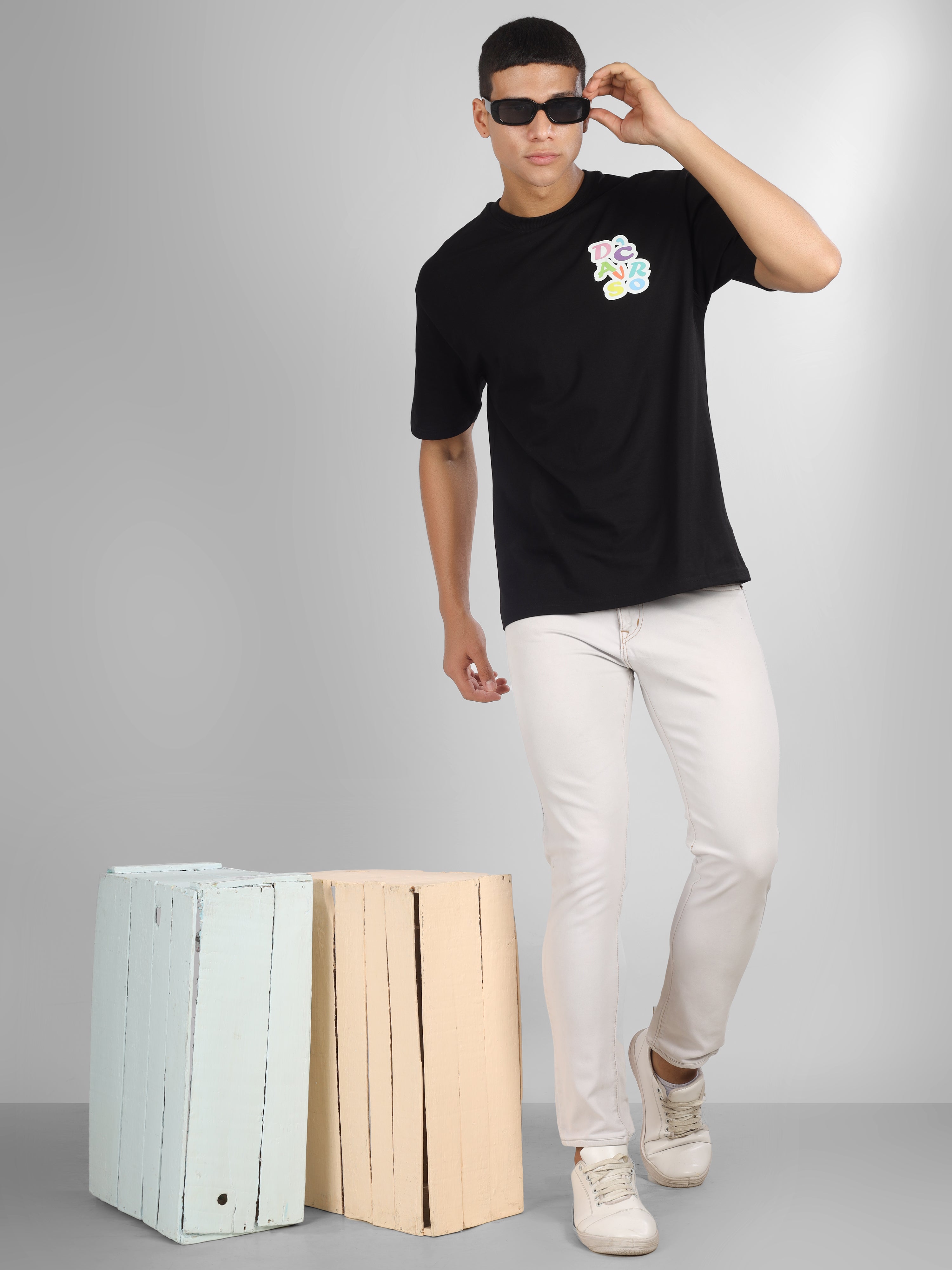 Male model wearing a black oversized dcavros T-shirt with a vibrant multicolor logo on the left chest, paired with white jeans.