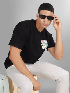 Male model wearing a black oversized dcavros T-shirt with a vibrant multicolor logo on the left chest, paired with white jeans.