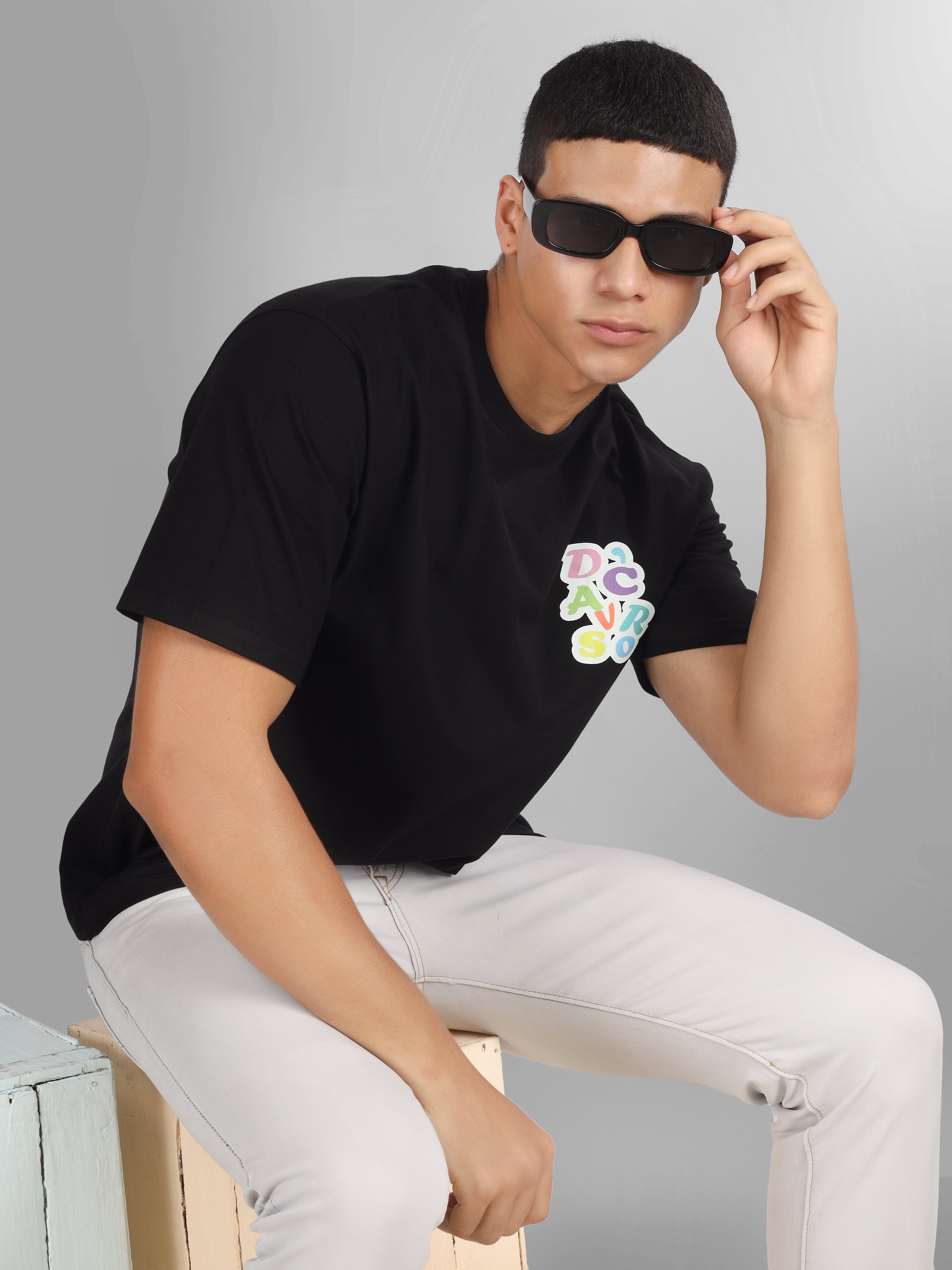 Male model wearing a black oversized dcavros T-shirt with a vibrant multicolor logo on the left chest, paired with white jeans.