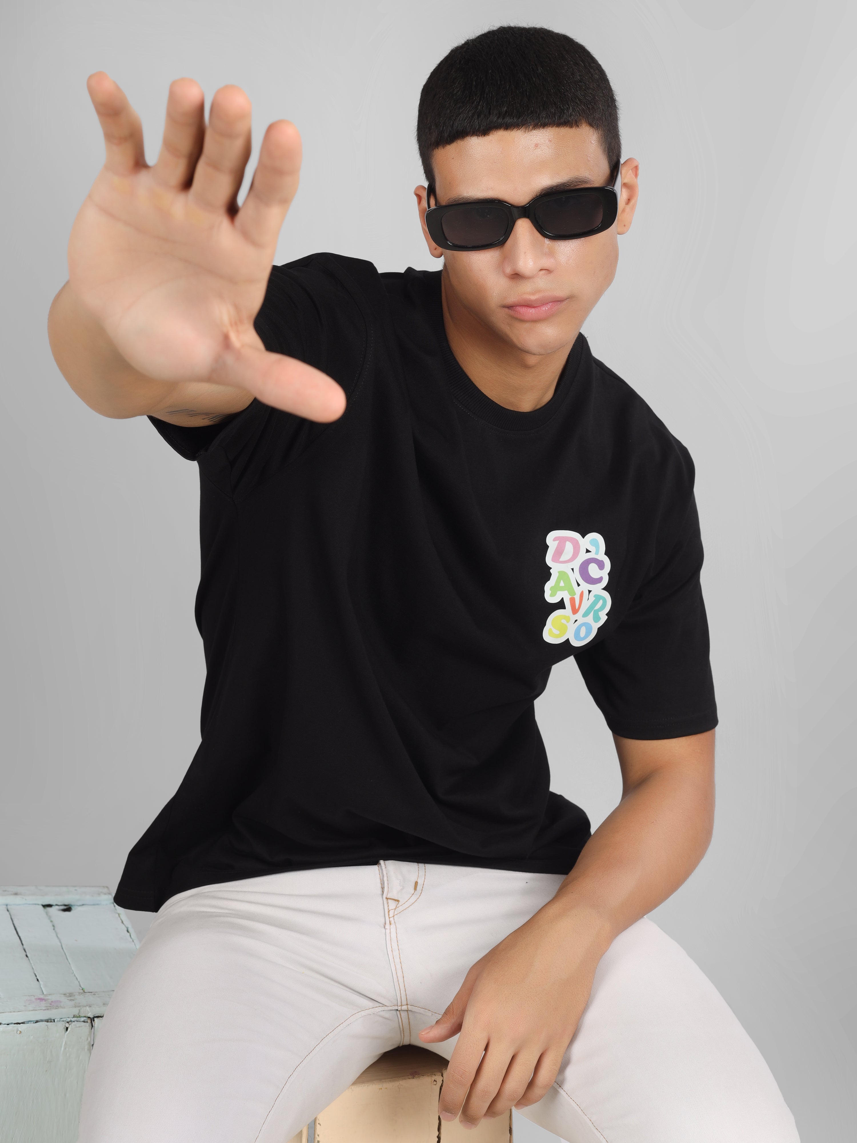 Male model wearing a black oversized dcavros T-shirt with a vibrant multicolor logo on the left chest, paired with white jeans.