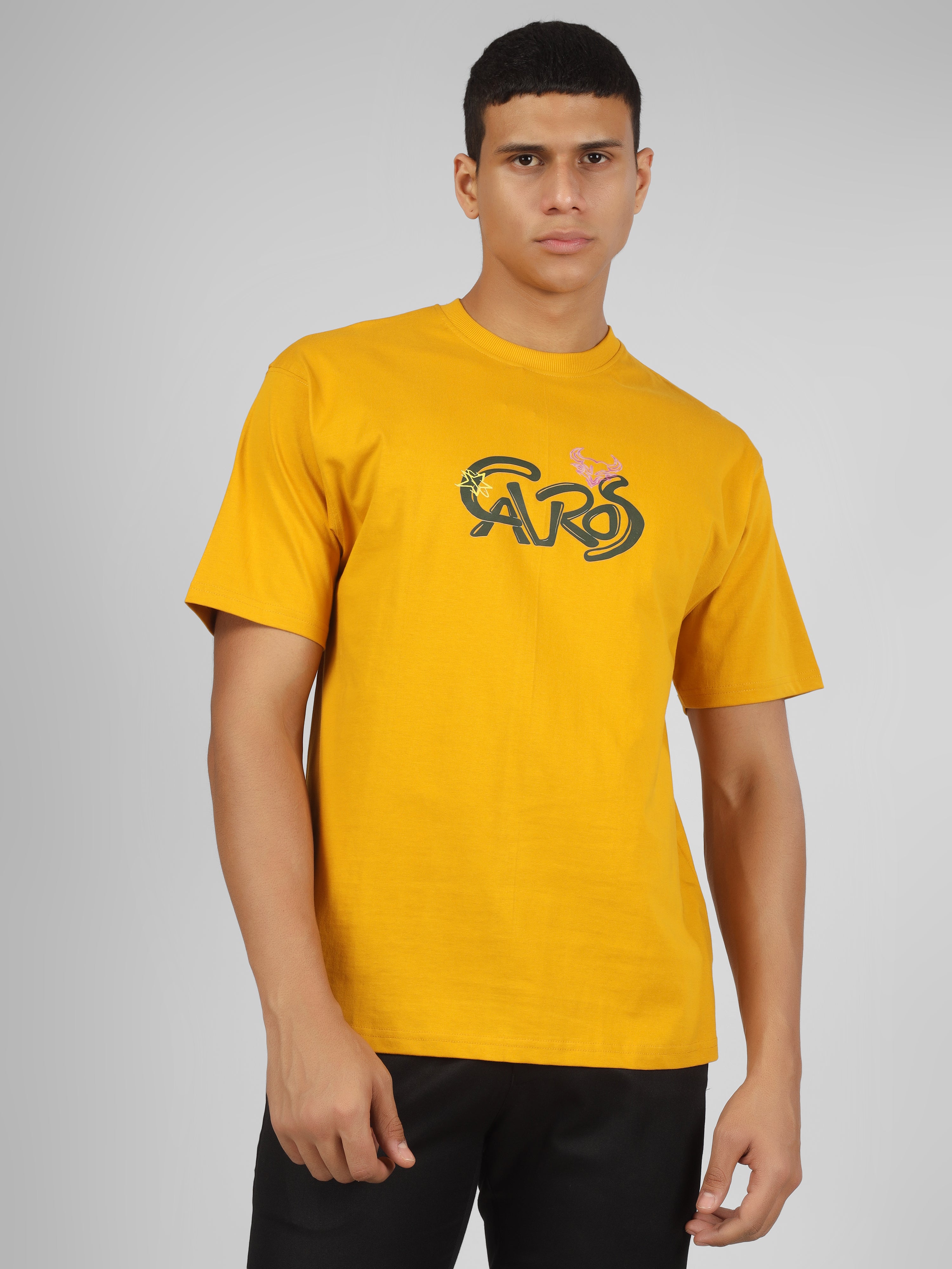 Male model wearing a mustard yellow dcavros unisex T-shirt featuring a bold “Cavros” front logo in black and pink, paired with black pants.