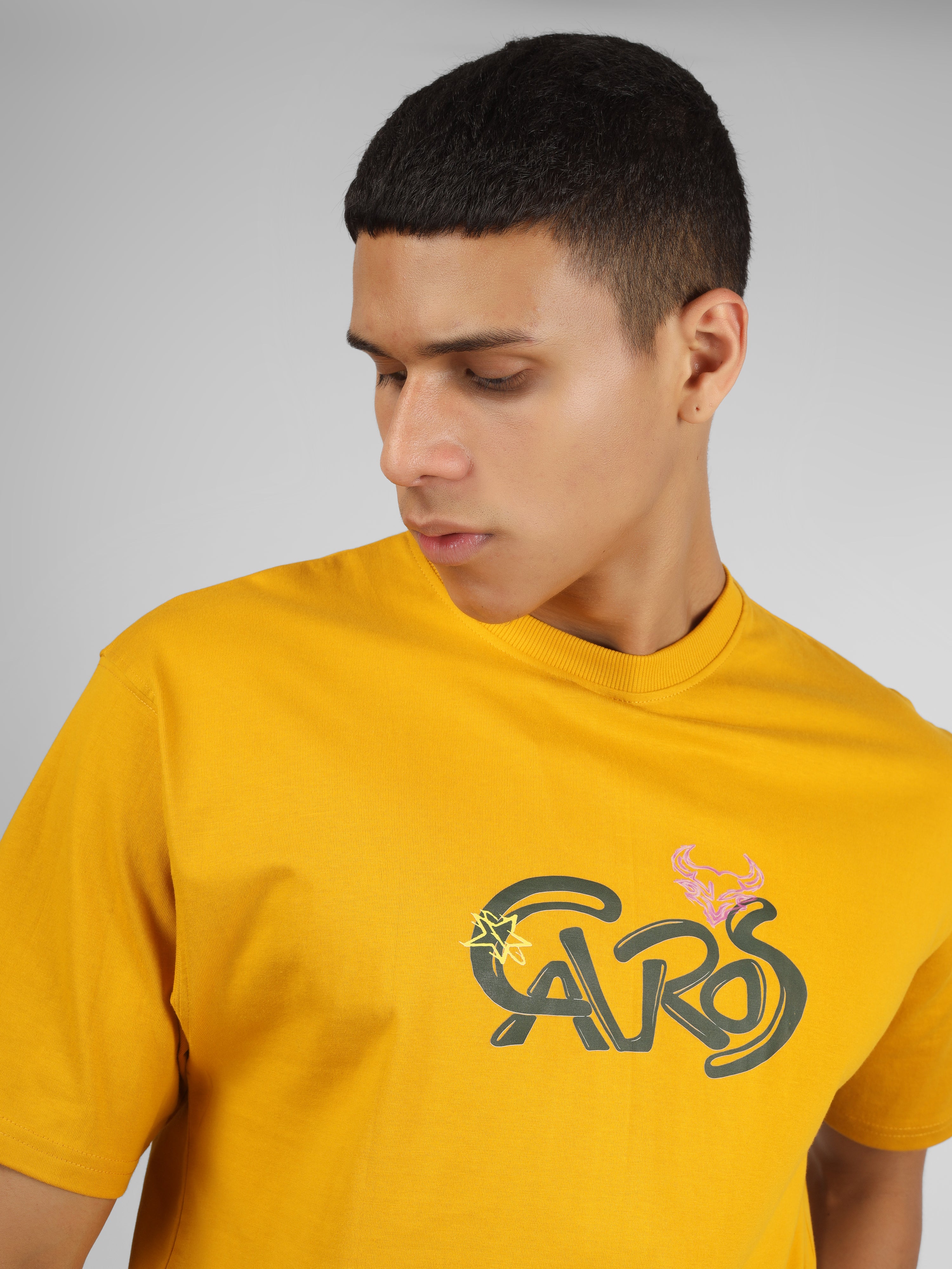Male model wearing a mustard yellow dcavros unisex T-shirt featuring a bold “Cavros” front logo in black and pink, paired with black pants.