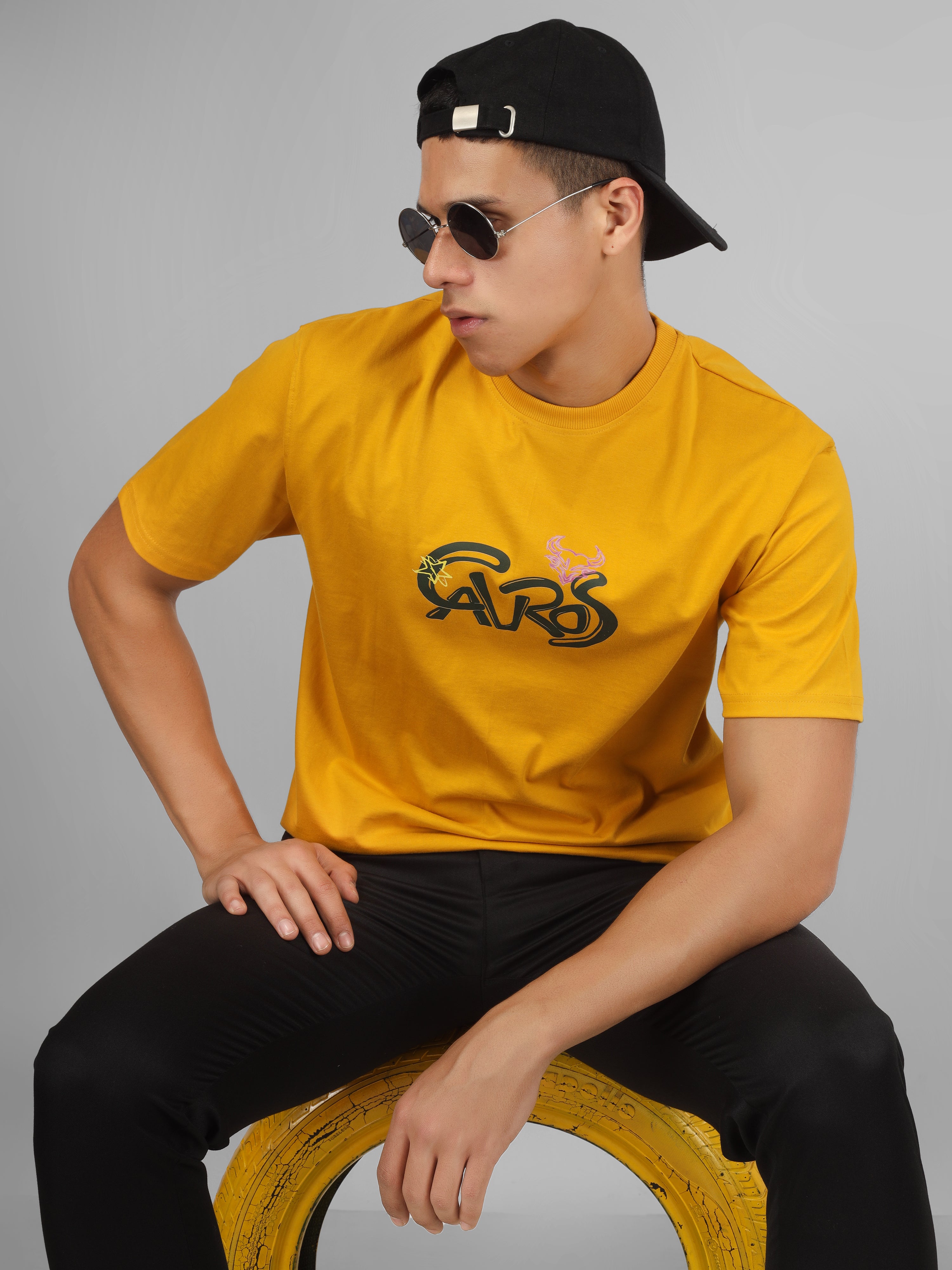 Male model wearing a mustard yellow dcavros unisex T-shirt featuring a bold “Cavros” front logo in black and pink, paired with black pants.