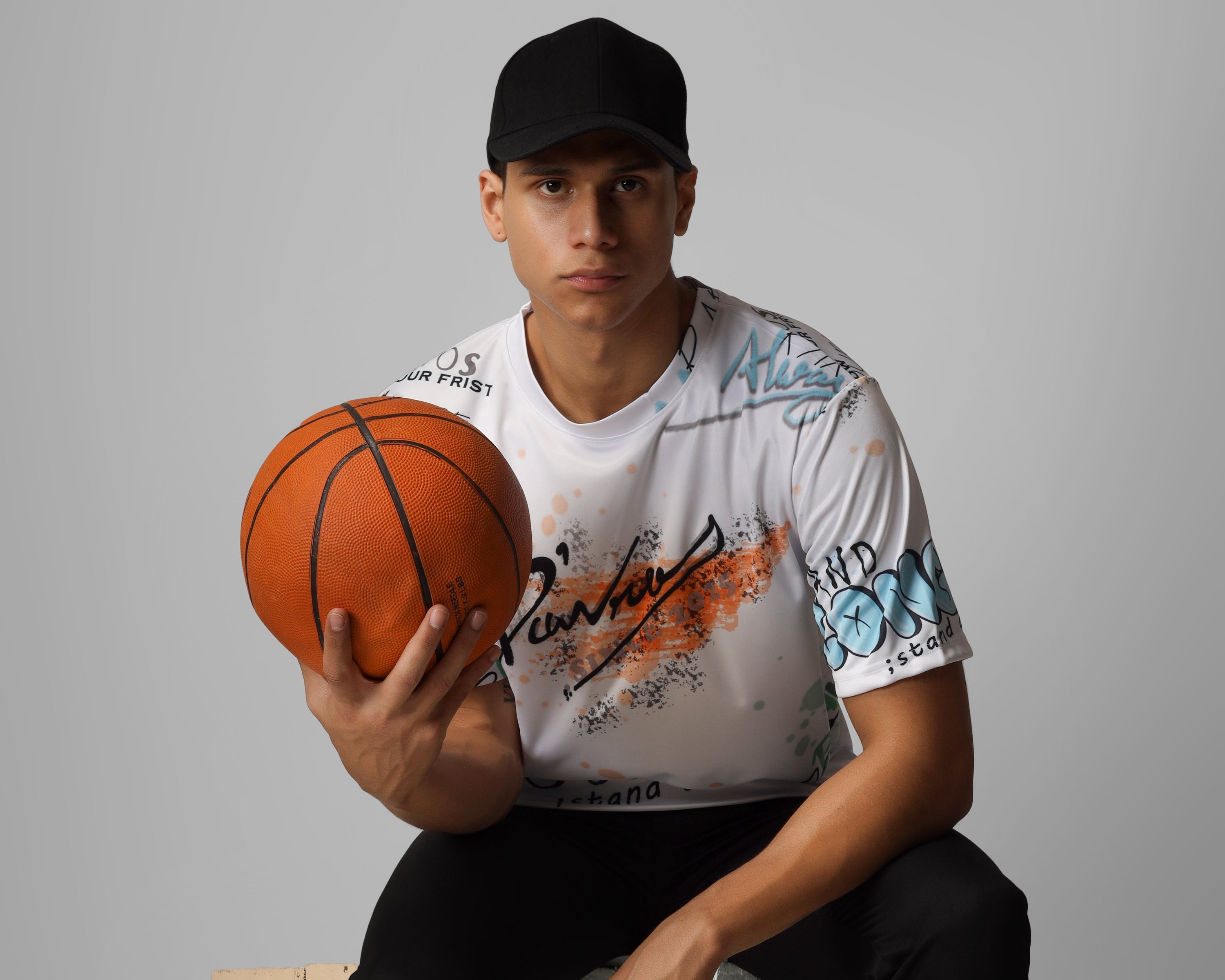 Person holding a basketball, wearing a white graphic dcavros t-shirt and black cap, sitting on a block with a gray background.