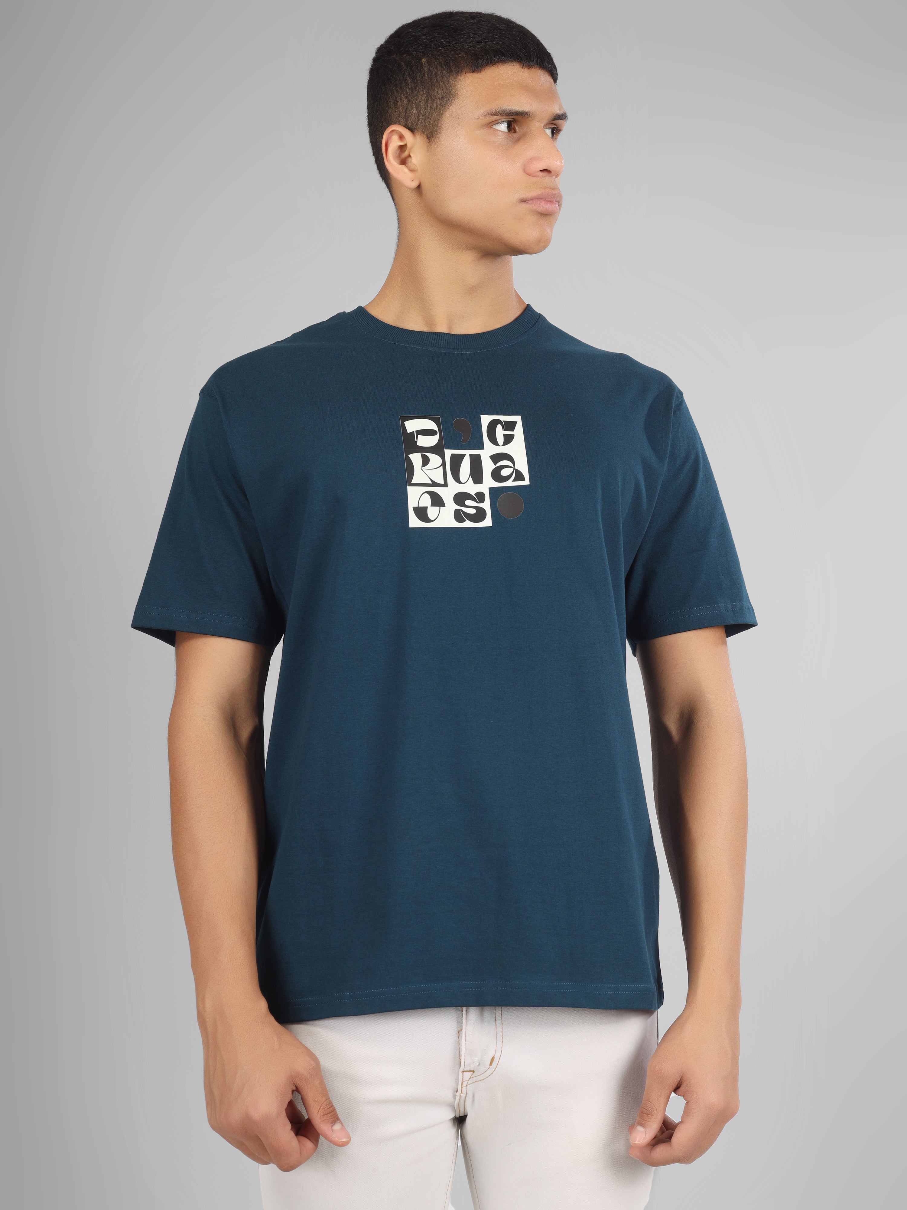Male model wearing a petrol blue dcavros T-shirt with a bold abstract central logo print in black and white, paired with white jeans.