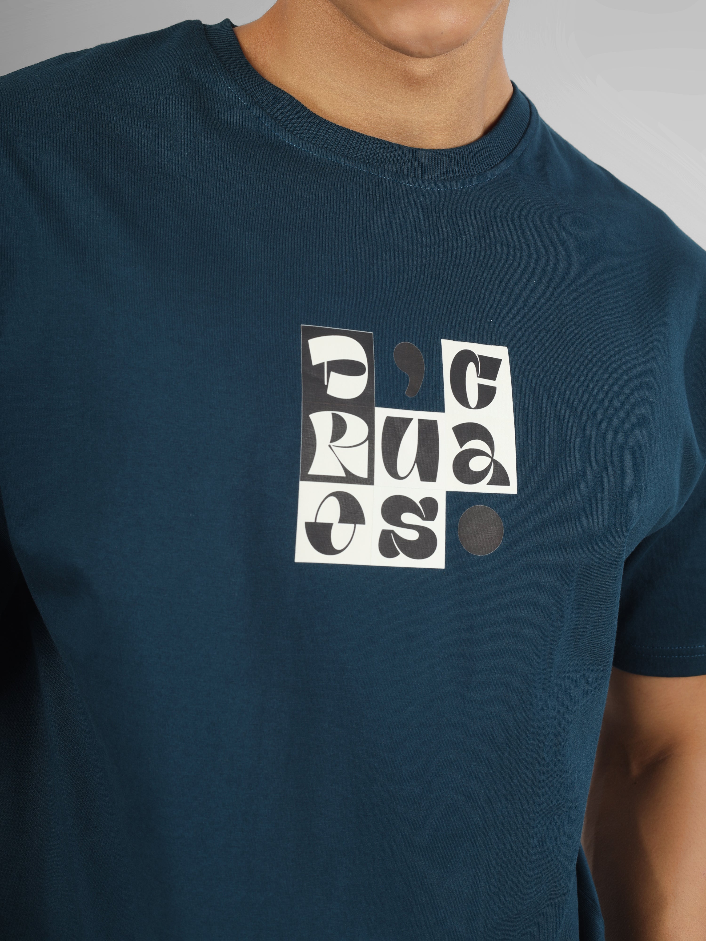 Male model wearing a petrol blue dcavros T-shirt with a bold abstract central logo print in black and white, paired with white jeans.