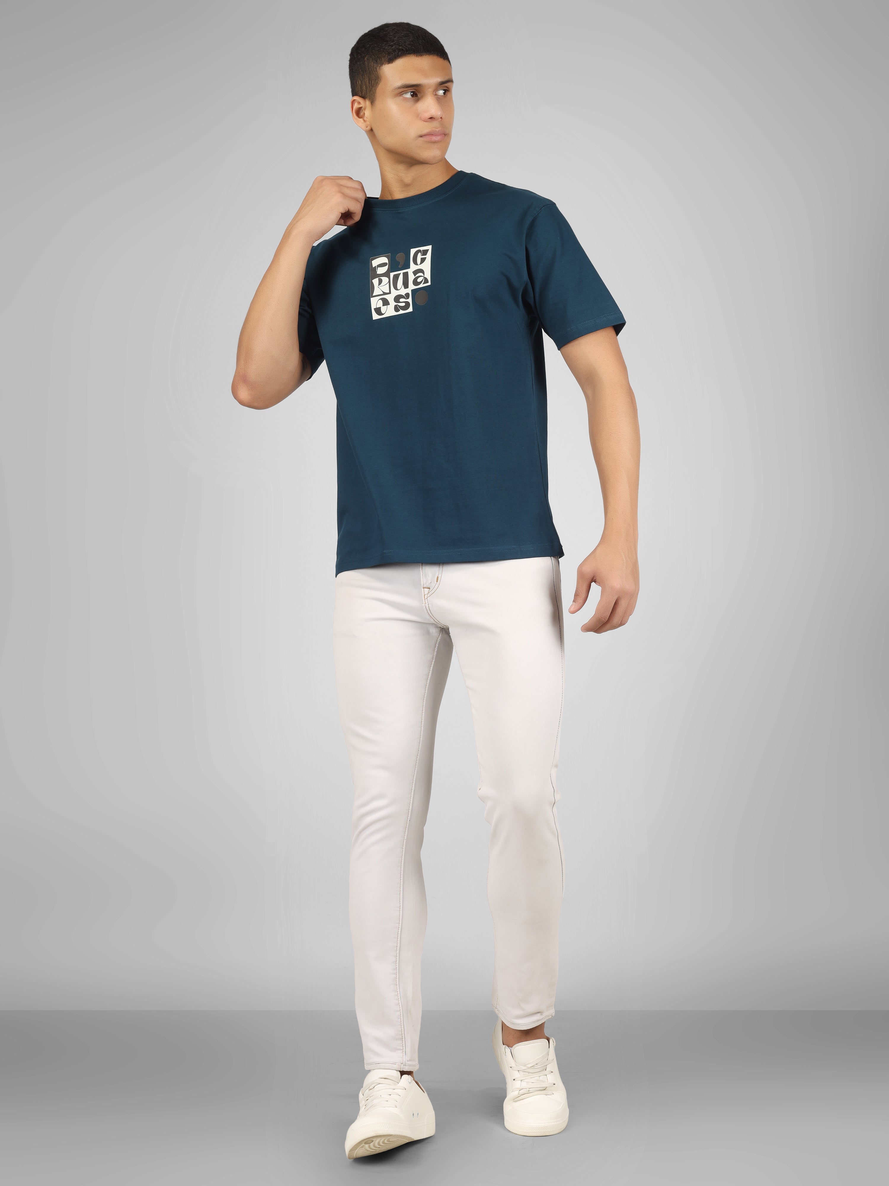 Male model wearing a petrol blue dcavros T-shirt with a bold abstract central logo print in black and white, paired with white jeans.