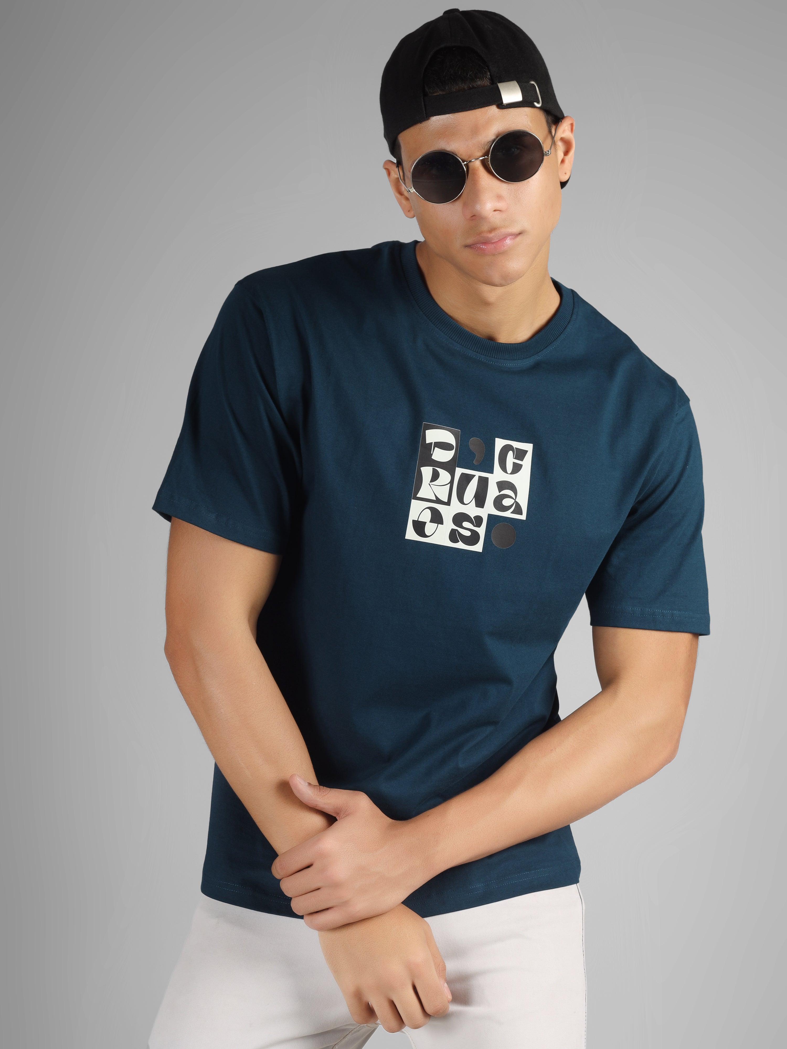 Male model wearing a petrol blue dcavros T-shirt with a bold abstract central logo print in black and white, paired with white jeans.