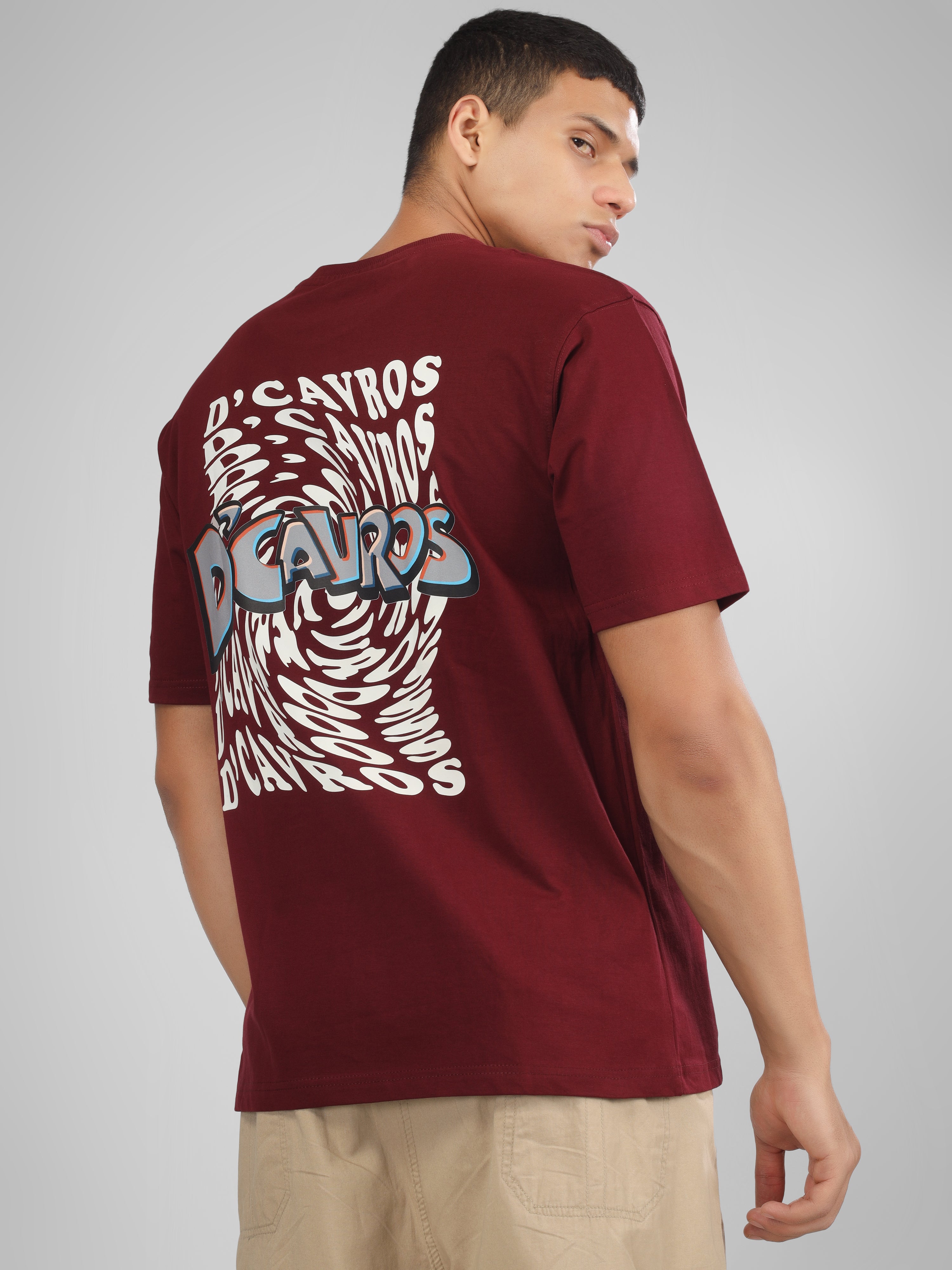 Male model wearing a maroon dcavros graphic T-shirt with a spiral text logo on the back and relaxed fit, styled with beige jeans.