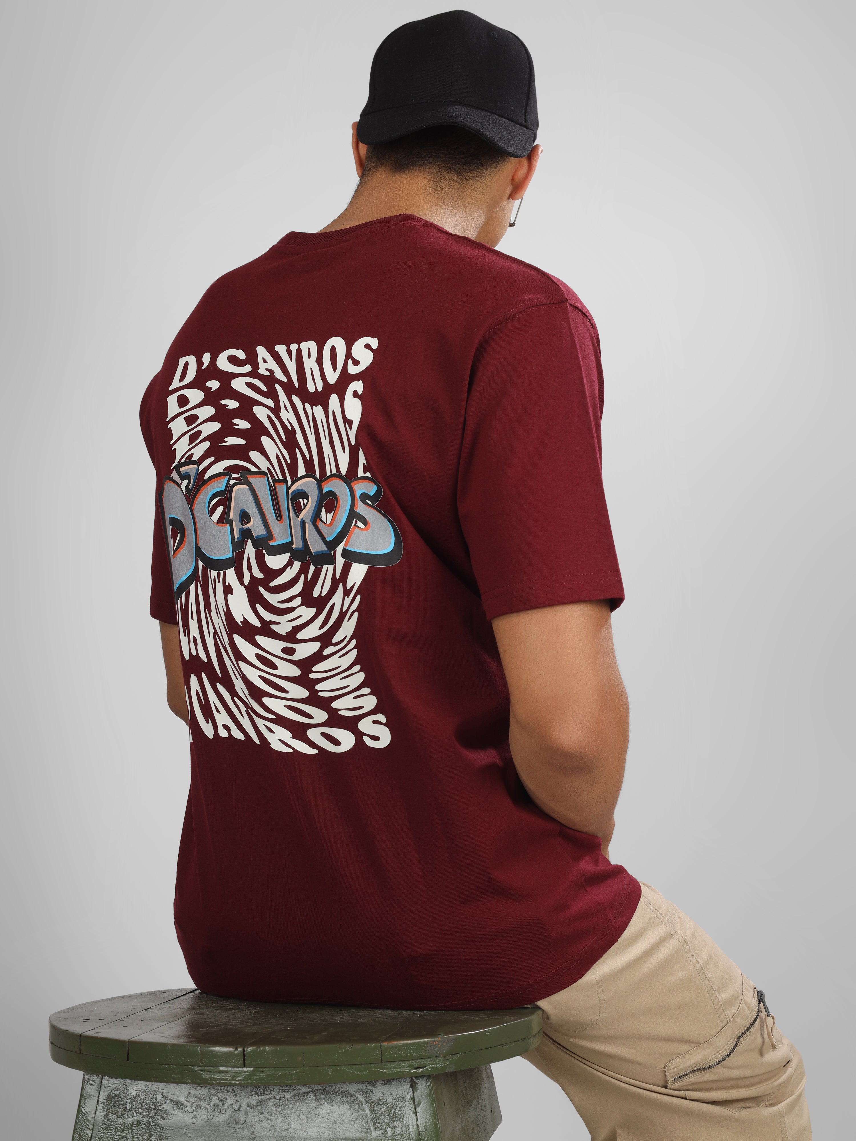 Male model wearing a maroon dcavros graphic T-shirt with a spiral text logo on the back and relaxed fit, styled with beige jeans.