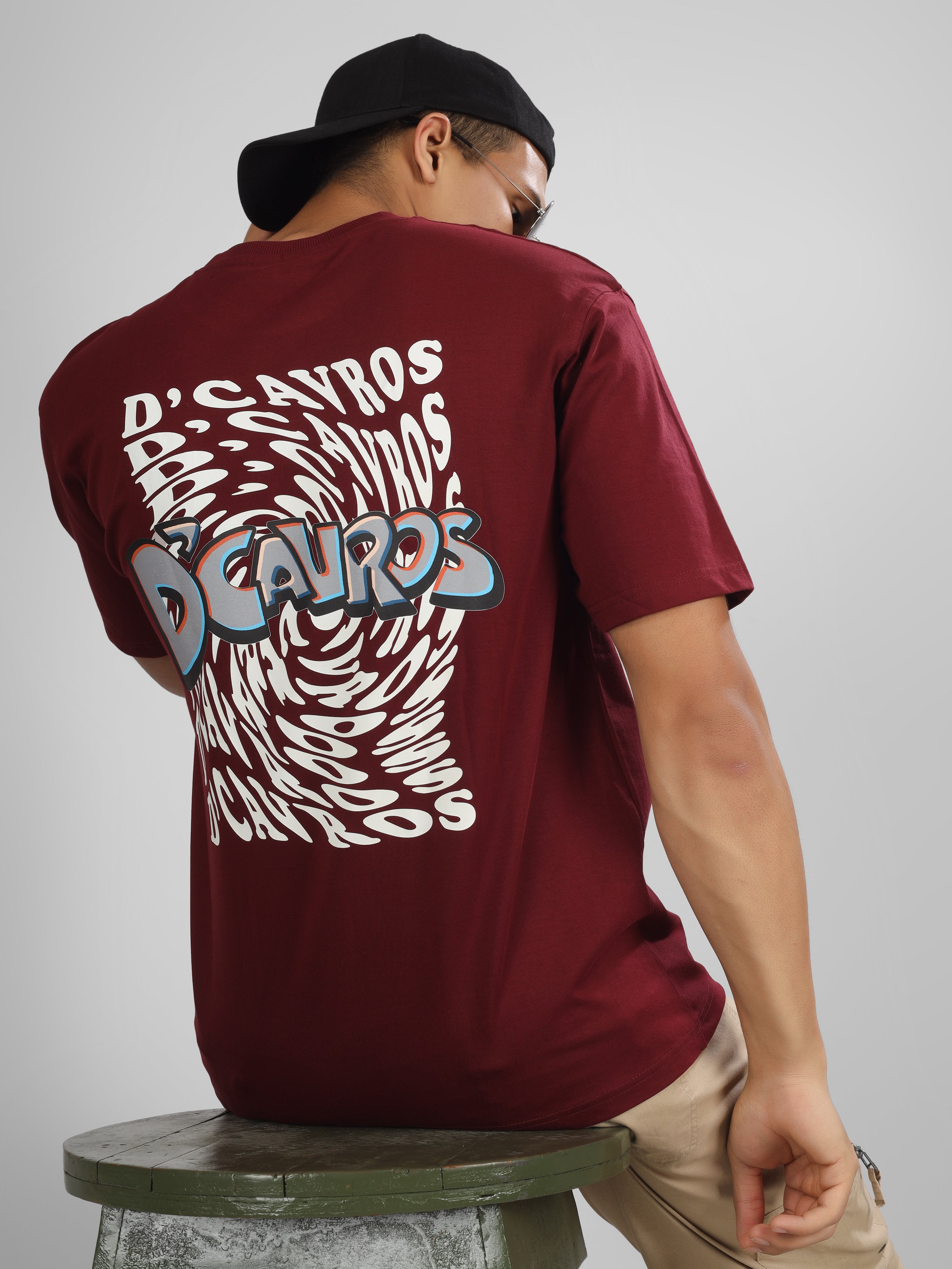 Male model wearing a maroon dcavros graphic T-shirt with a spiral text logo on the back and relaxed fit, styled with beige jeans.
