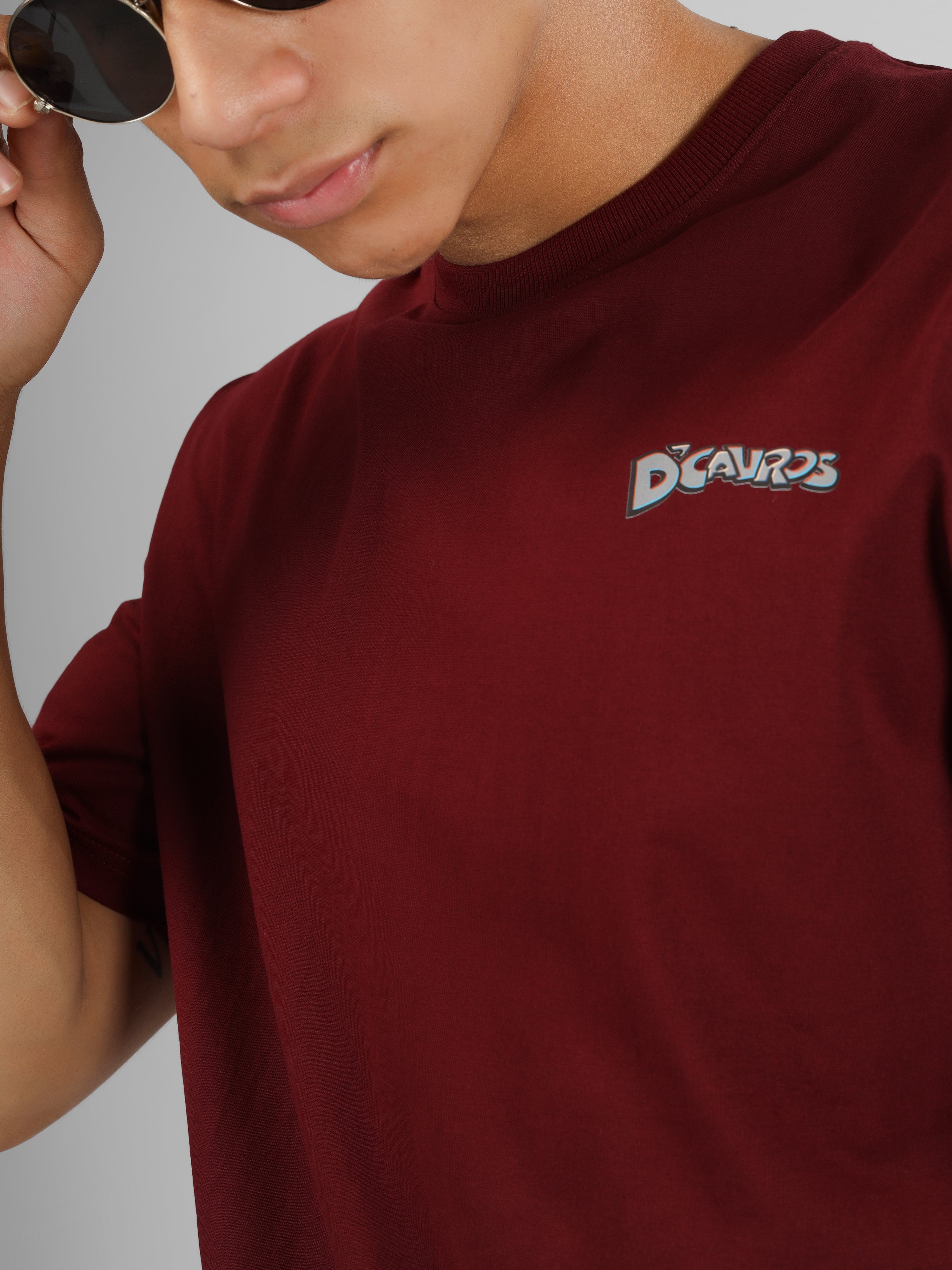 Male model wearing a maroon dcavros graphic T-shirt with a spiral text logo on the back and relaxed fit, styled with beige jeans.