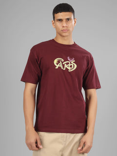 Male model wearing a deep maroon dcavros unisex T-shirt with a stylized yellow and pink "Cavros" bull logo on the front, paired with beige pants.