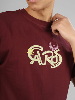 Male model wearing a deep maroon dcavros unisex T-shirt with a stylized yellow and pink "Cavros" bull logo on the front, paired with beige pants.
