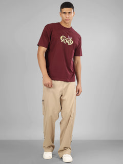 Male model wearing a deep maroon dcavros unisex T-shirt with a stylized yellow and pink "Cavros" bull logo on the front, paired with beige pants.