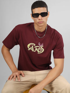 Male model wearing a deep maroon dcavros unisex T-shirt with a stylized yellow and pink "Cavros" bull logo on the front, paired with beige pants.