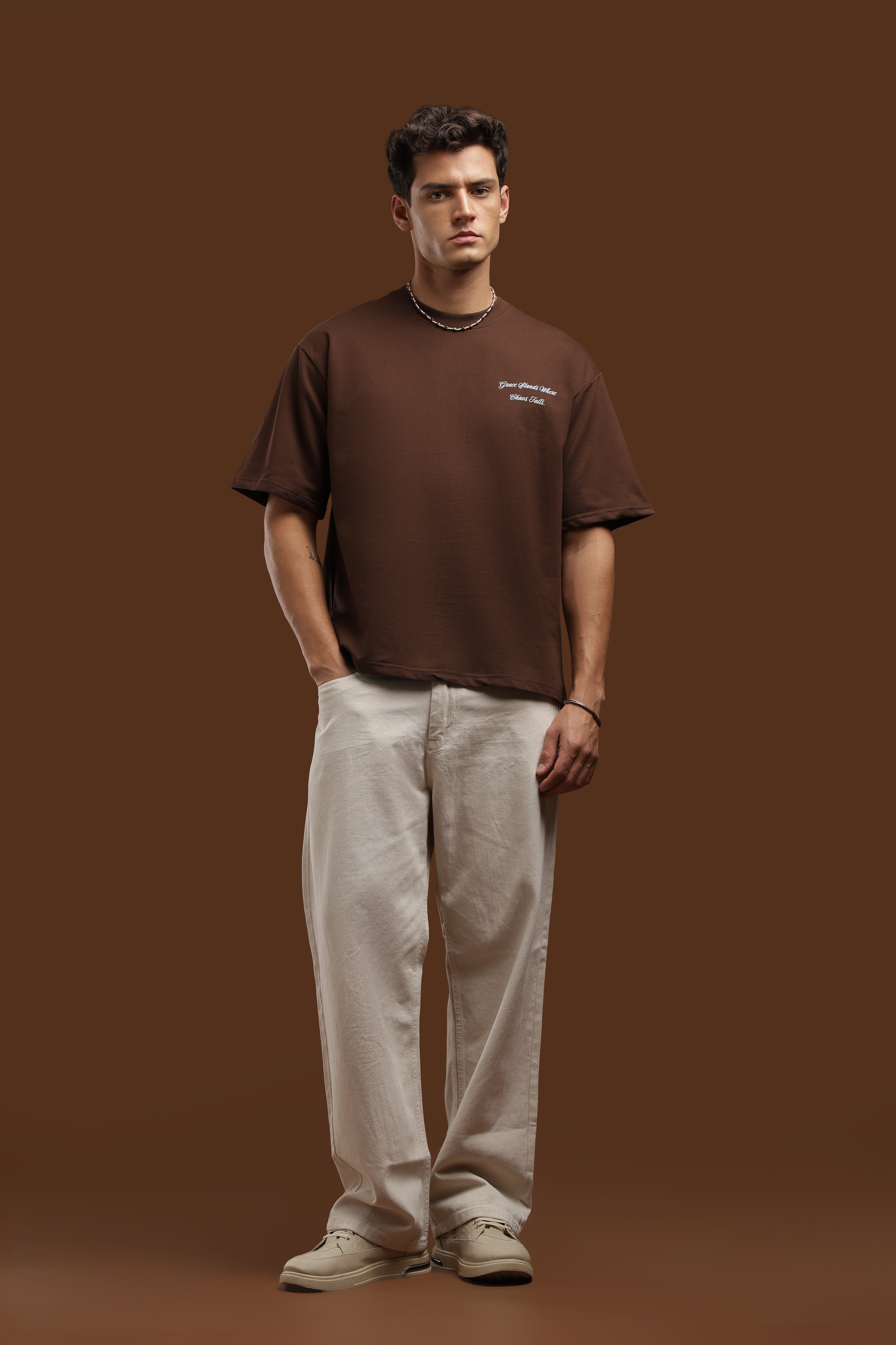 Model wearing an Oversized Fit Graphics Printed T-Shirt in brown, paired with light pants and a stylish necklace.