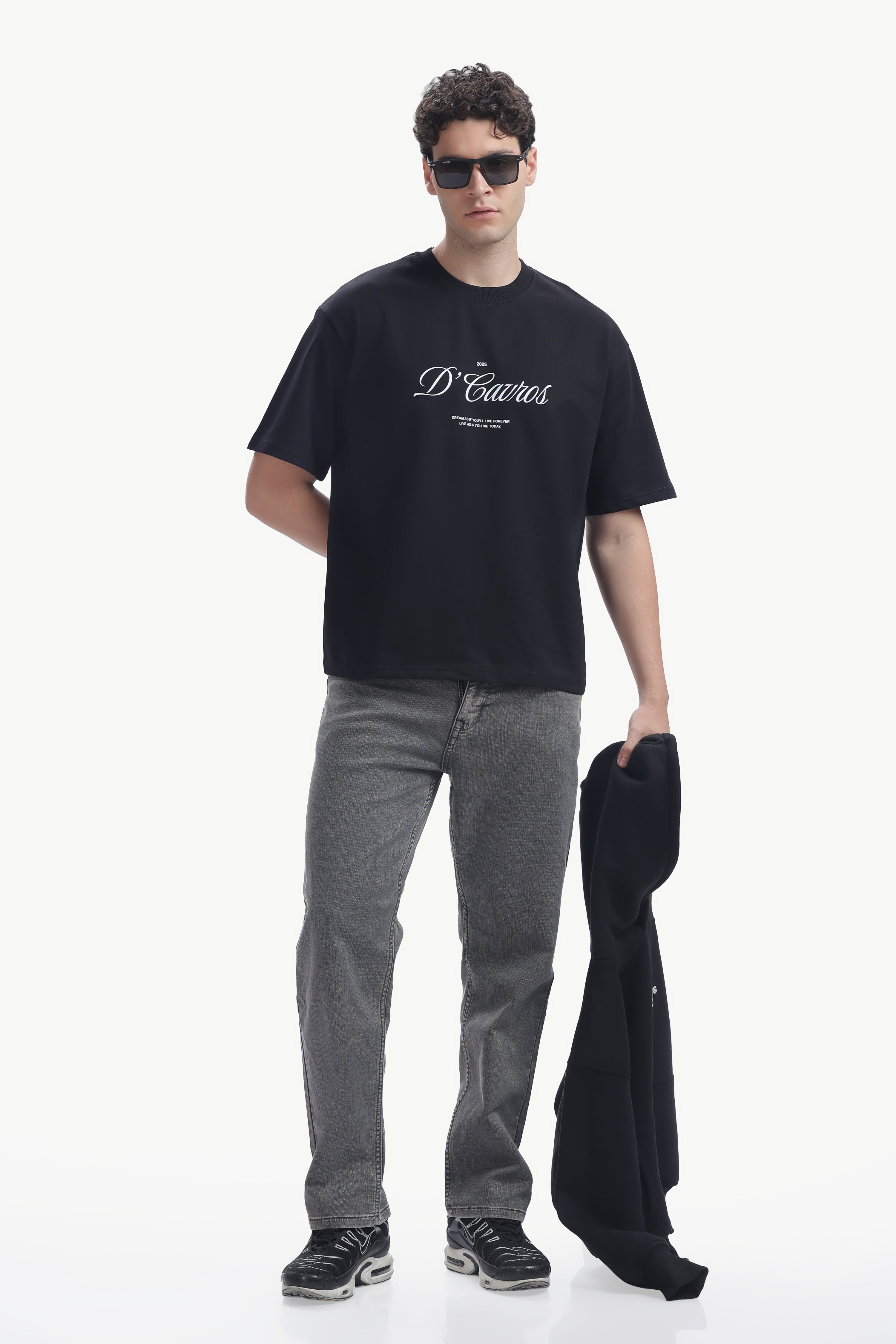Model wearing a Classic Loose Fit Typography T-Shirt in black with D'Cavros script, paired with grey jeans and sunglasses.