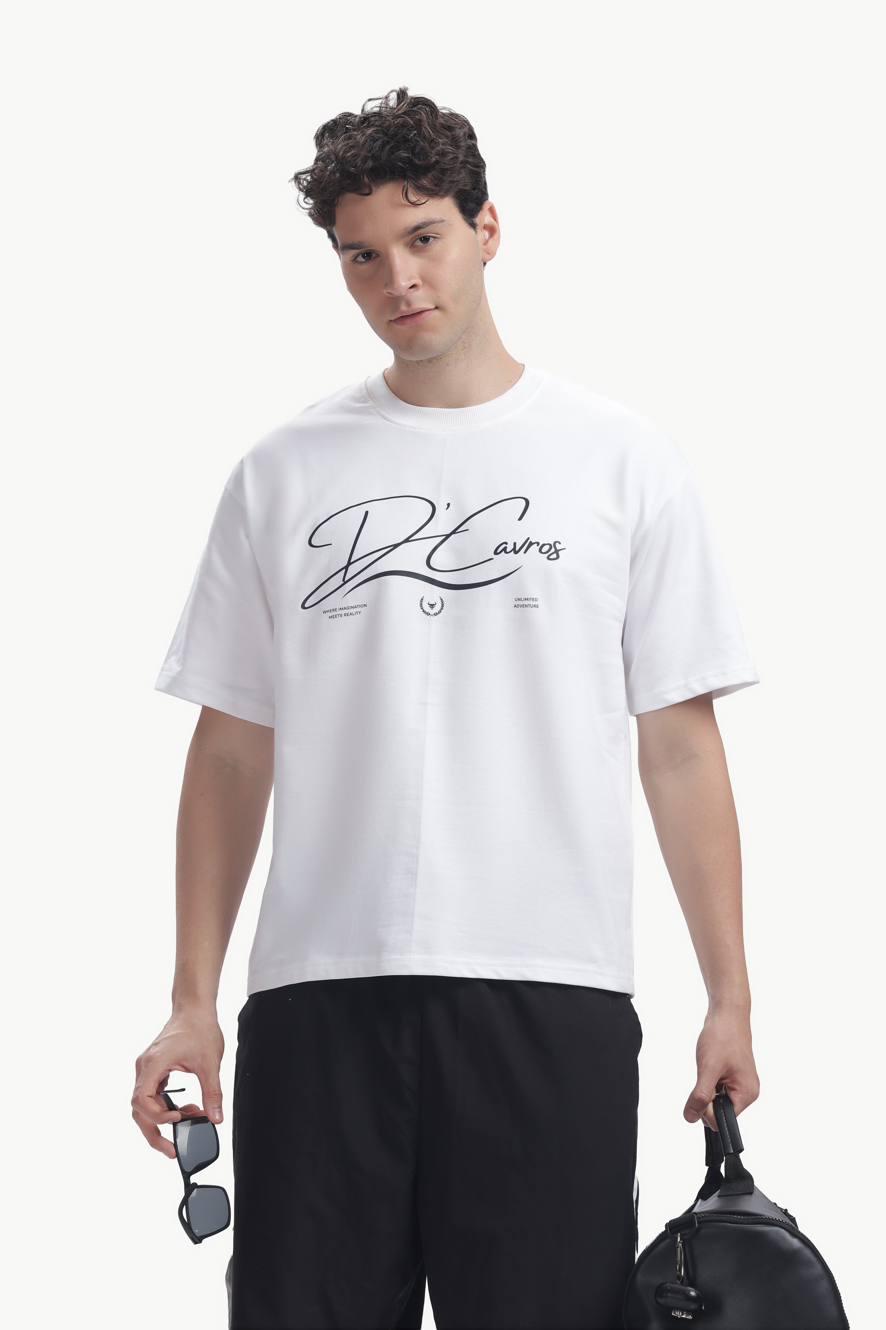 Man wearing Community Capsule Graphics Tee in white with bold typography, sunglasses in hand, and a stylish bag.