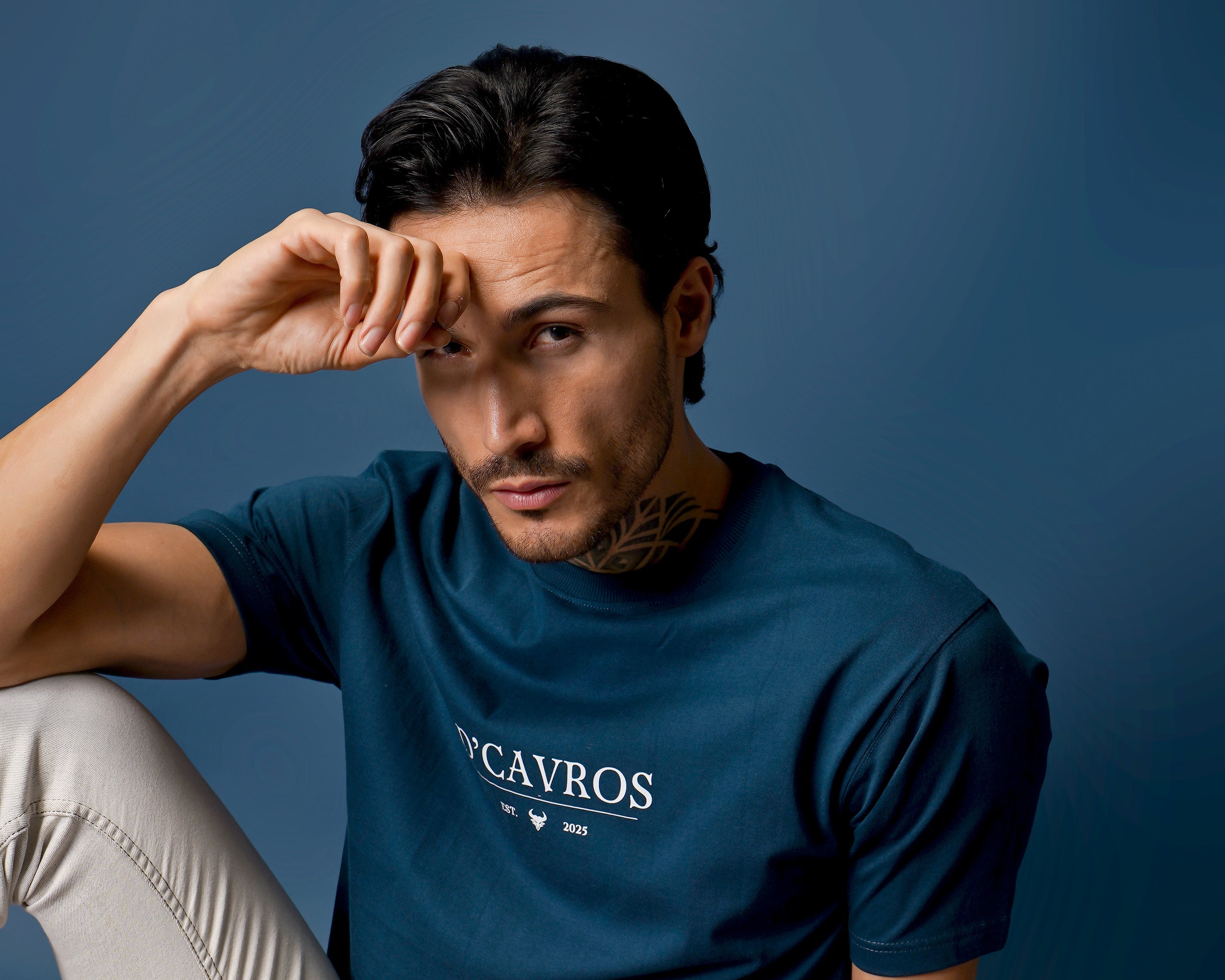 Man wearing a blue t-shirt with 'D'Cavros' logo sitting on a colorful rug against a dark blue wall.