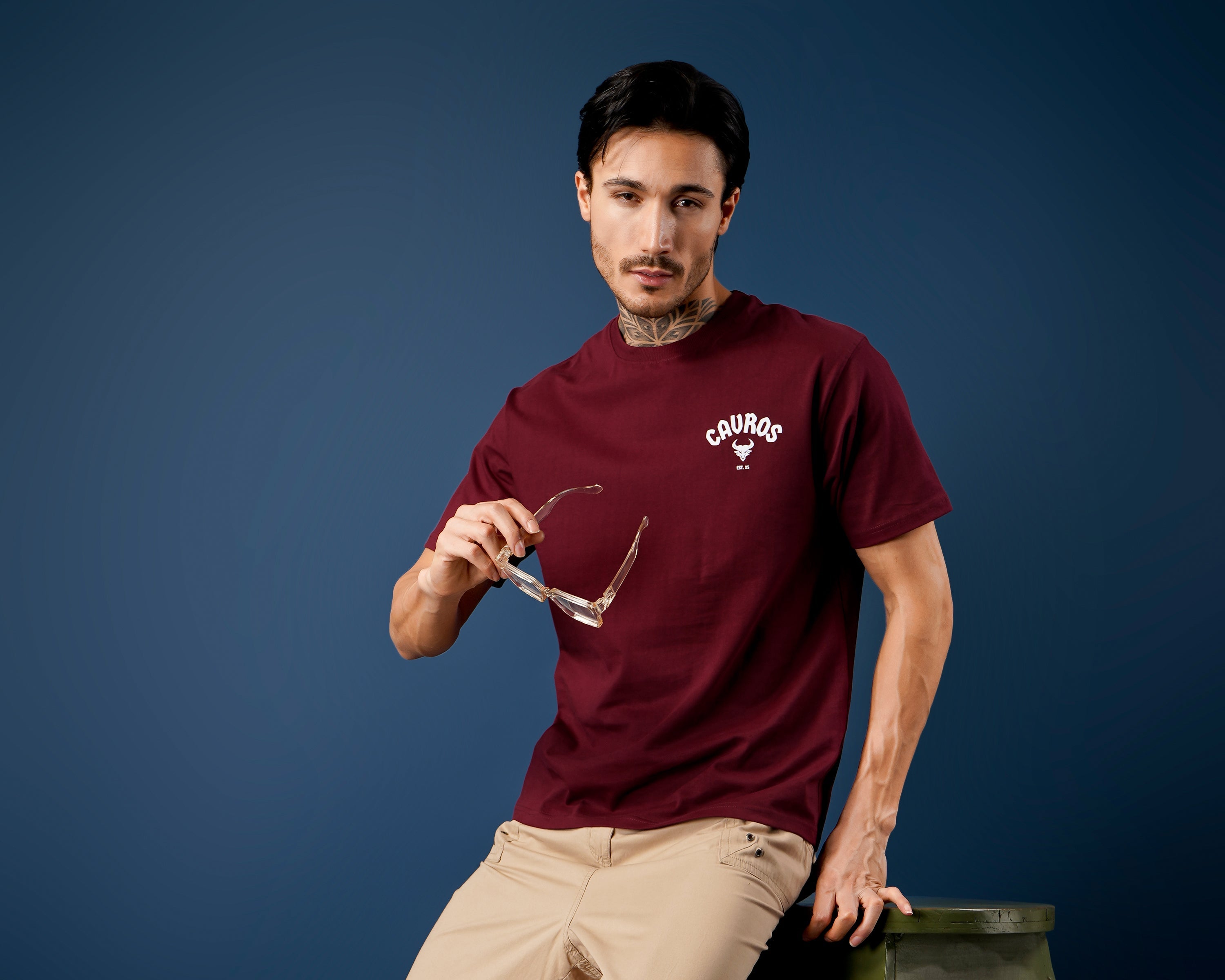 Man wearing a maroon dcavros t-shirt and beige pants standing on a colorful rug with a dark blue background