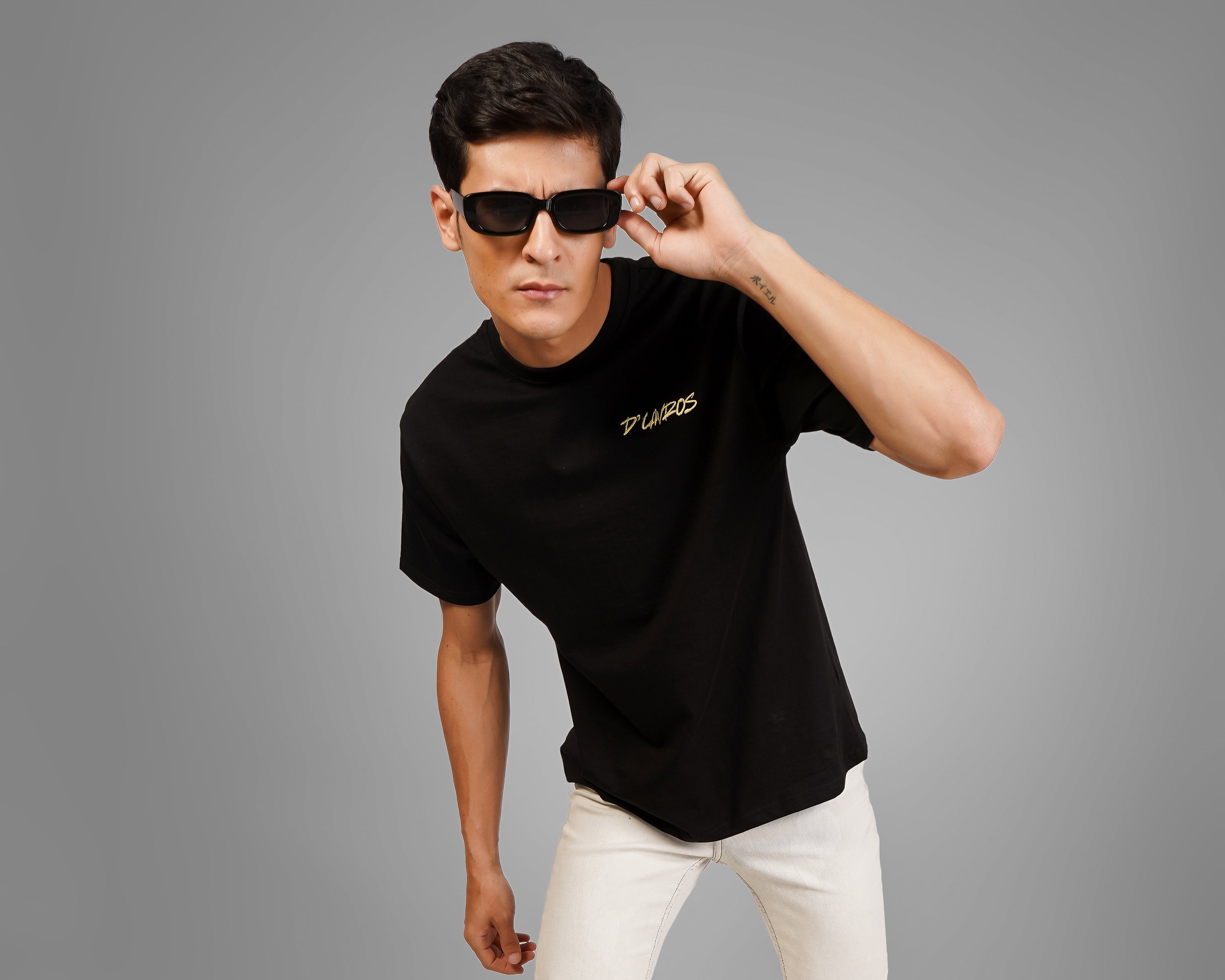 Man wearing a black dcavros branding  t-shirt, white pants, and sunglasses on a gray background
